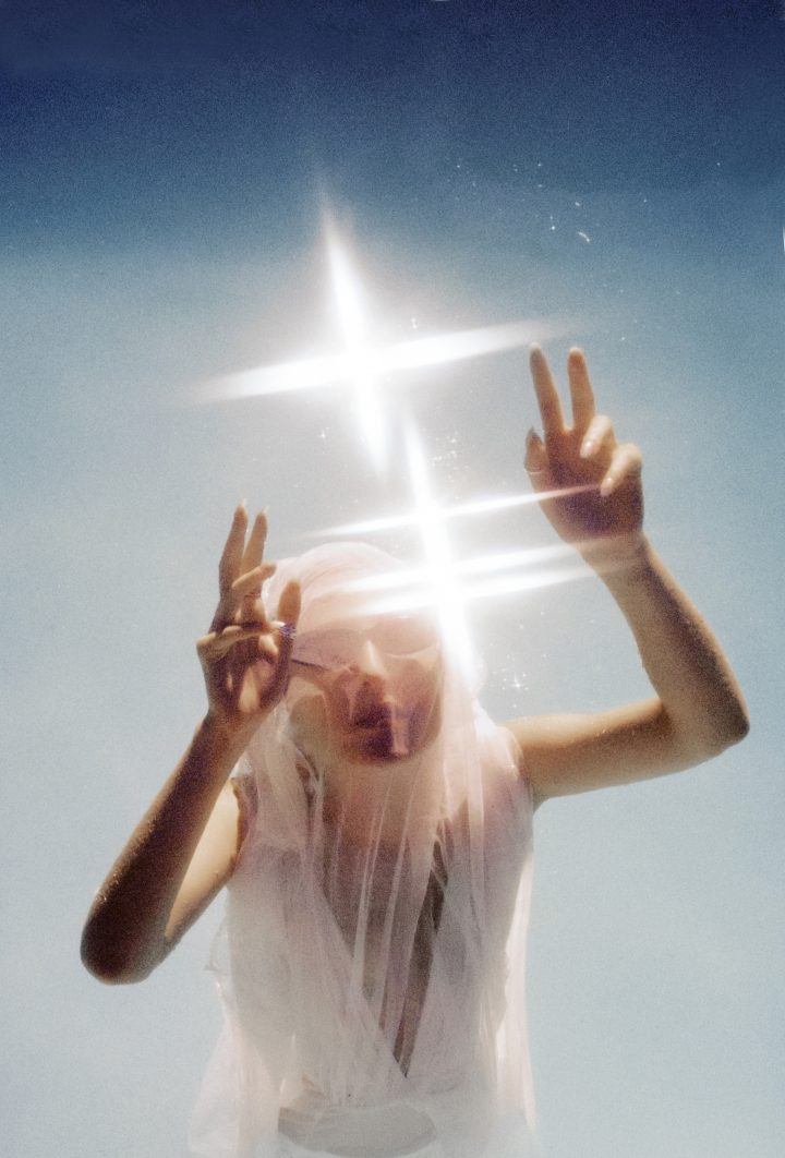 Selected works by Dimitri Guedes — A figure in translucent white fabric is photographed from a low angle against a gradient sky, with a bright cross-shaped light source obscuring the head and...