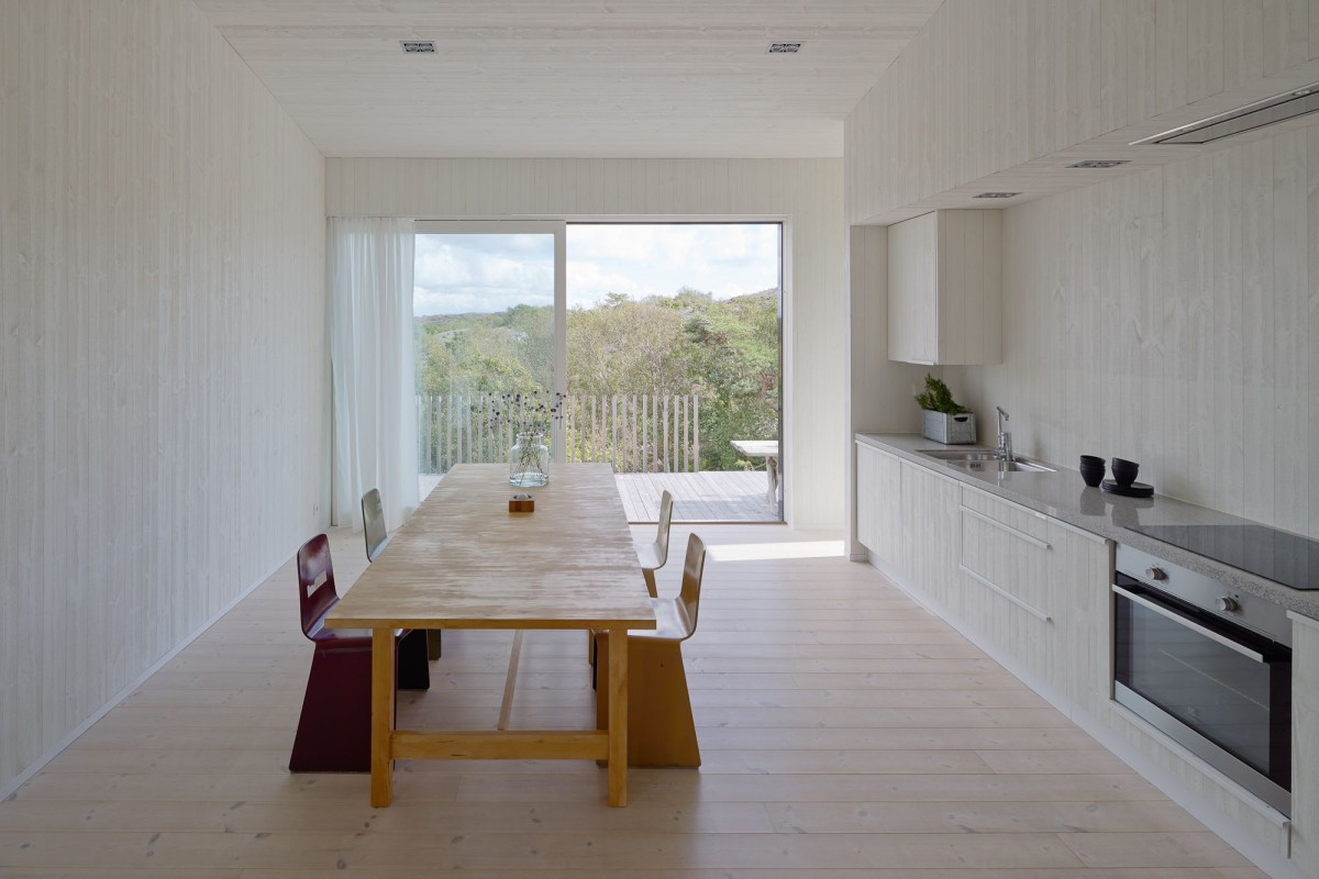 Kyrkesund Weekend House by Mattias Gunneflo — A timber-floored kitchen and dining space extends toward a sliding glass door framing forested landscape.