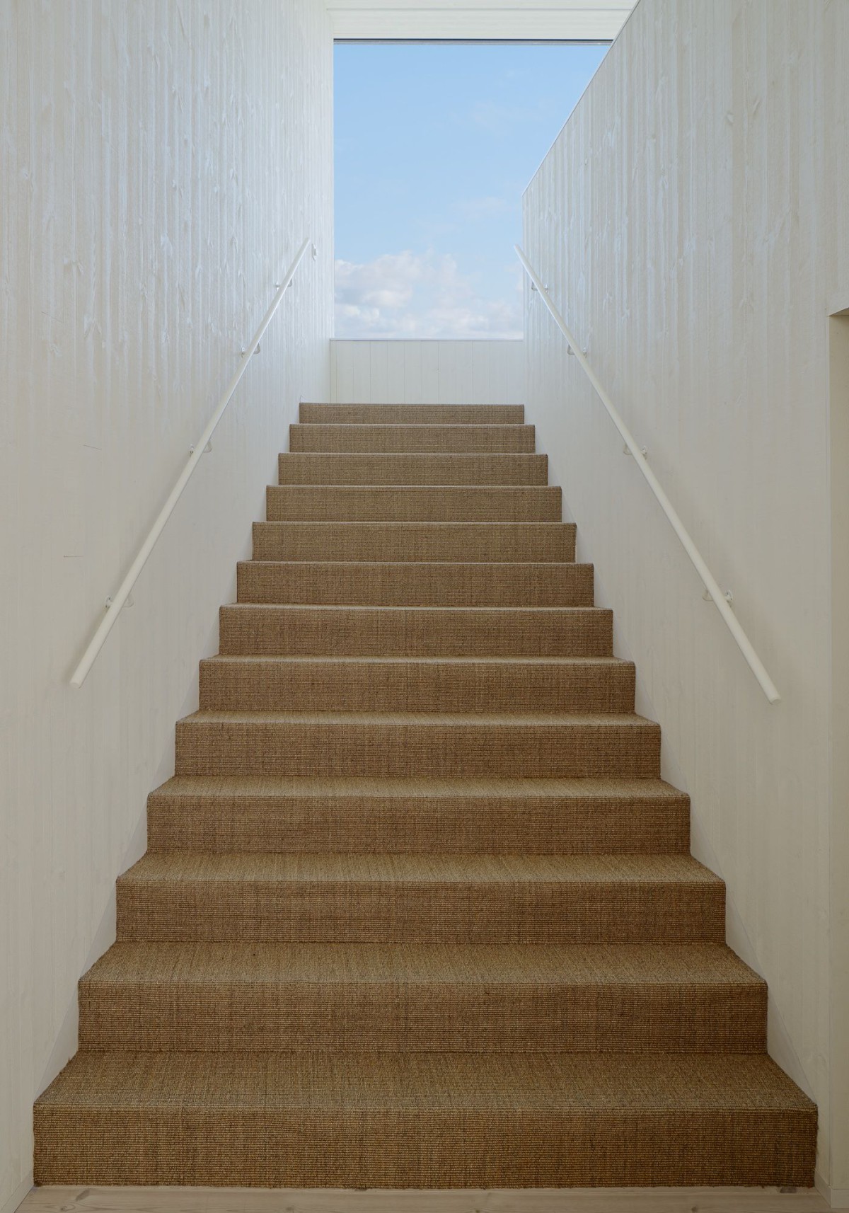 Kyrkesund Weekend House by Mattias Gunneflo — A stairwell ascends through a narrow concrete passage toward daylight, its walls rendered in off-white plaster forming a stark vertical corridor.