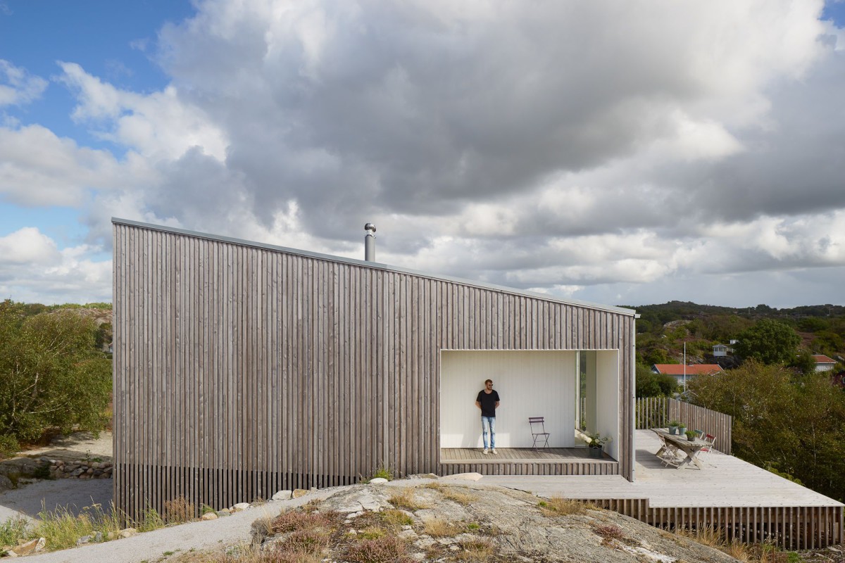 Kyrkesund Weekend House by Mattias Gunneflo — A corrugated metal-clad weekend house elevated on timber supports sits on exposed bedrock, its weathered steel facade punctuated by a horizontal ribbon window.