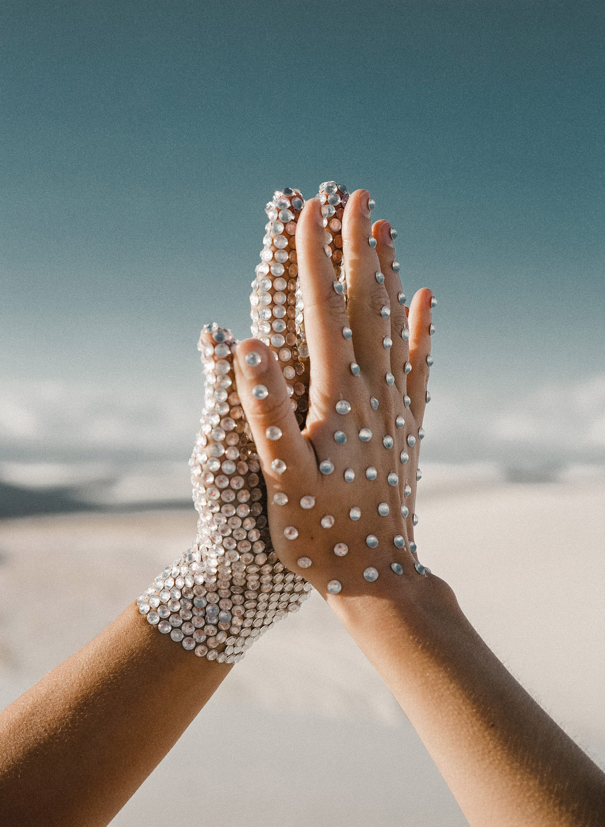 Selected works by Leeor Wild — Two hands raised against a teal sky and distant beach, both adorned with pearl-like spherical beads covering the skin in dense, uniform patterns.