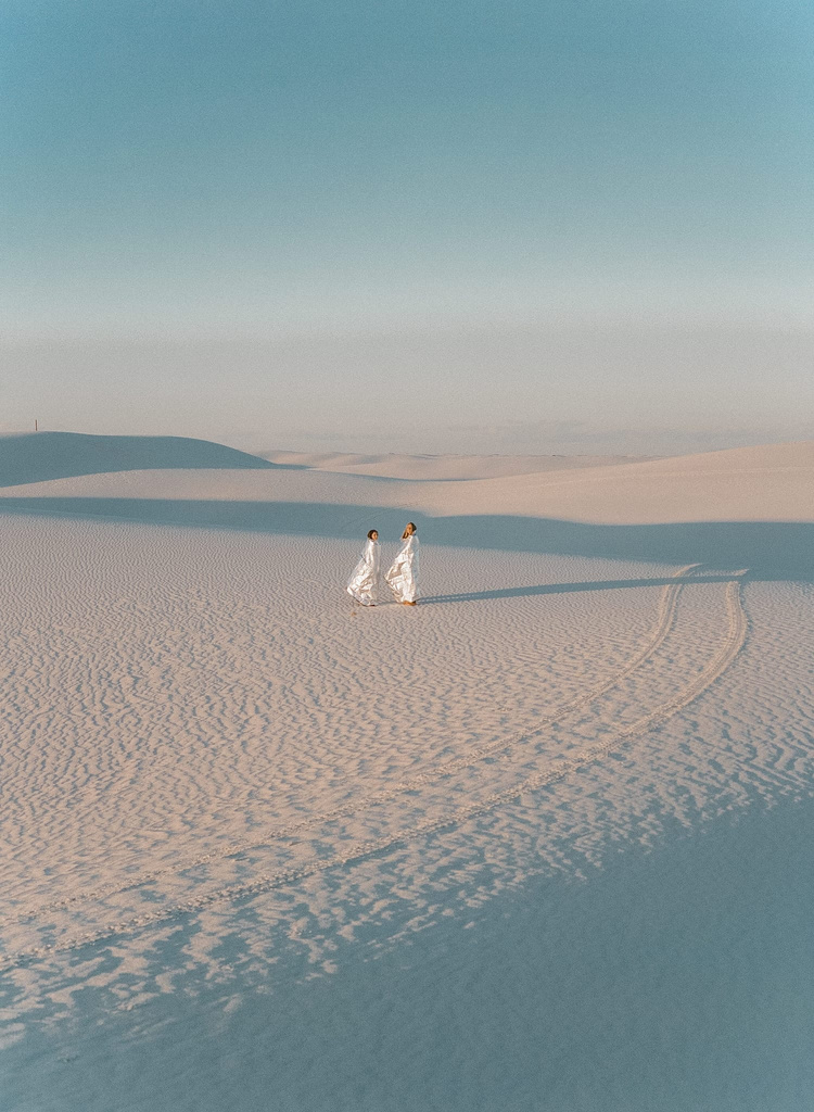 Selected works by Leeor Wild — Two figures in white linen garments traverse a vast desert landscape, their elongated shadows cast across rippled sand formations.