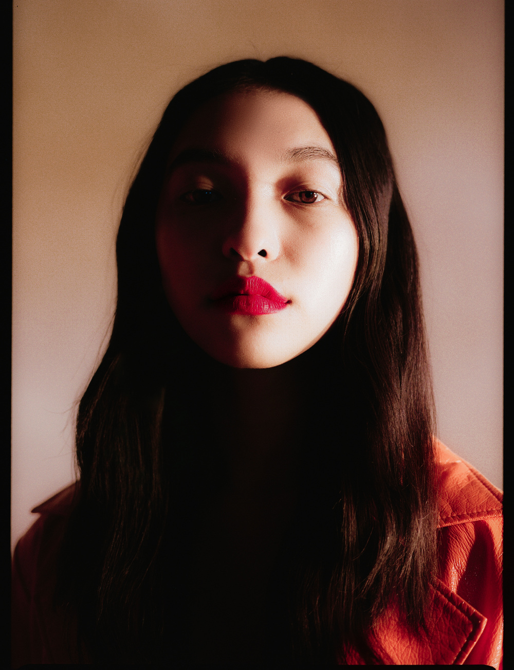Selected works by Leeor Wild — A portrait photograph centered on a figure with long dark hair and vivid crimson lipstick, positioned against a warm beige wall.