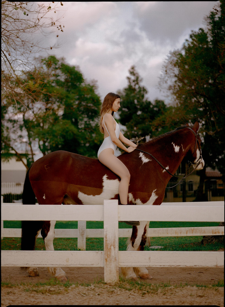 Selected works by Leeor Wild — A woman in a white swimsuit sits bareback on a brown and white horse positioned behind a white wooden ranch fence.