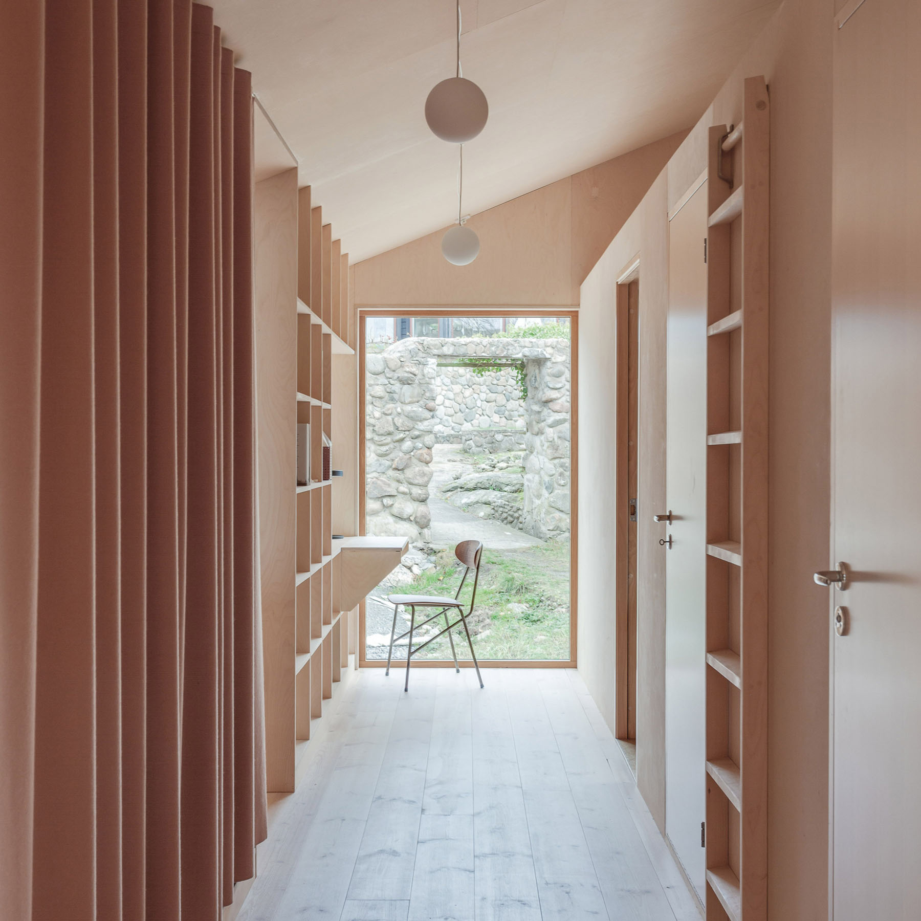 Villa Vassdal by Studio Holmberg — A corridor extends toward a framed threshold, where a solitary chair sits in the exterior courtyard beyond raw stone walls.