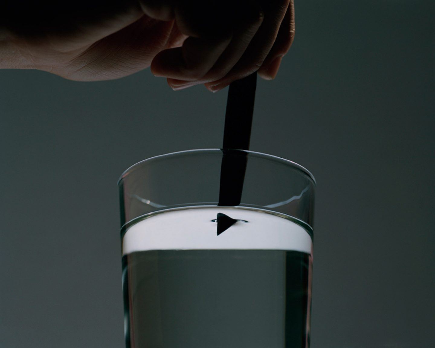 The Circle and the Line by Erik Östensson — A hand holds a black rod or stick submerged in a glass cylinder containing clear liquid with a white foam or cream layer at the surface.