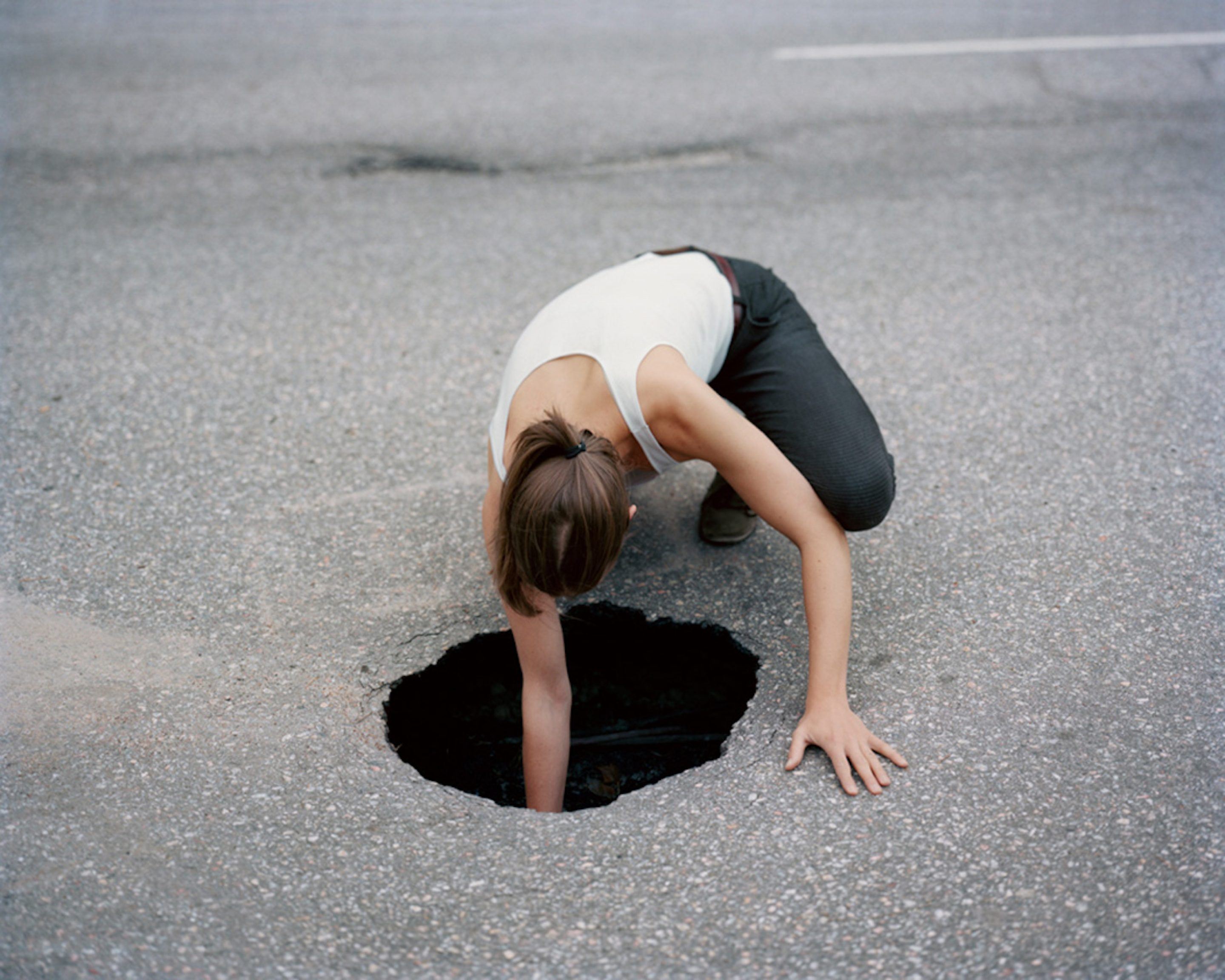 The Circle and the Line by Erik Östensson — A person in a white tank top and dark shorts bends forward into a near-complete fold, head tucked toward knees, positioned over a dark circular void in grey...