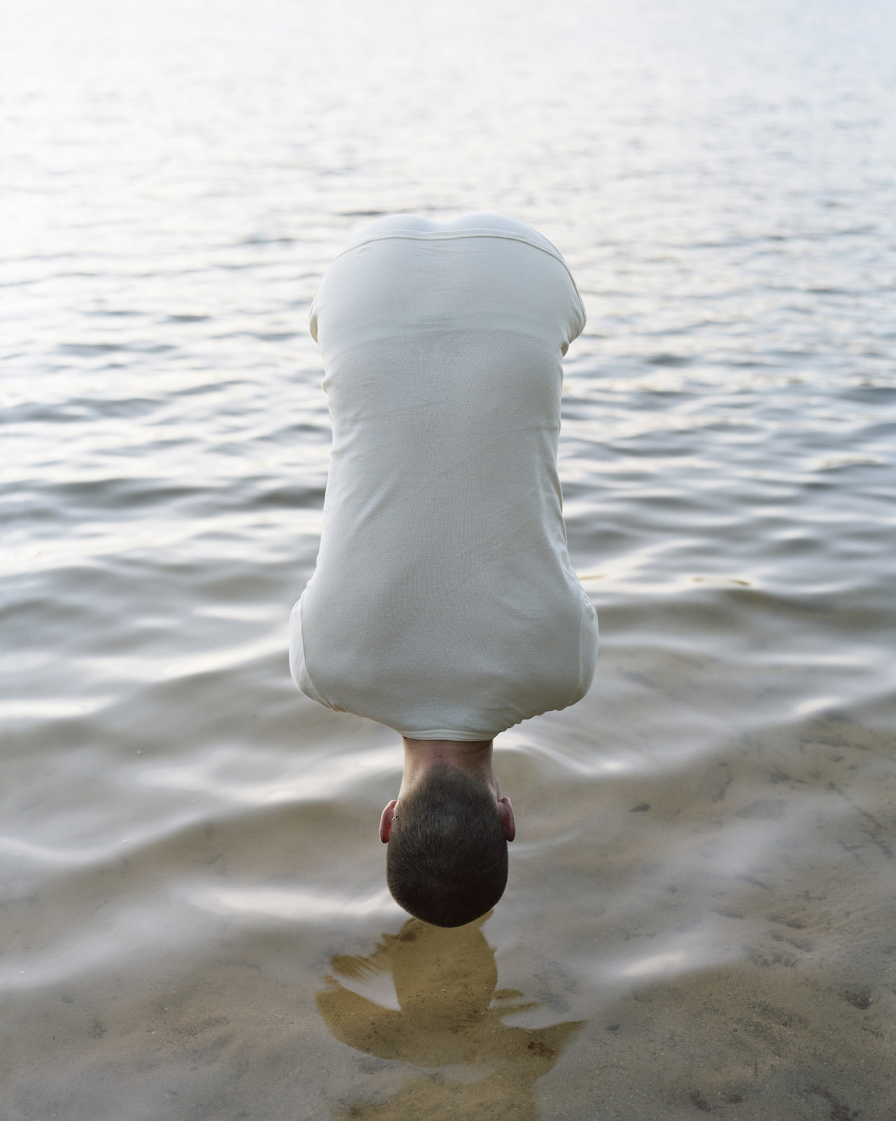 The Circle and the Line by Erik Östensson — A figure in a white linen shirt performs a headstand in shallow water, their dark hair visible at the apex while the garment creates a billowing,...