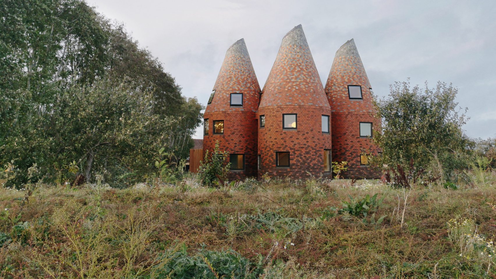 Bumpers Oast by ACME — Three conical brick towers with pitched roofs dominate a Kent landscape, their tapered forms arranged with visible gaps between structures.