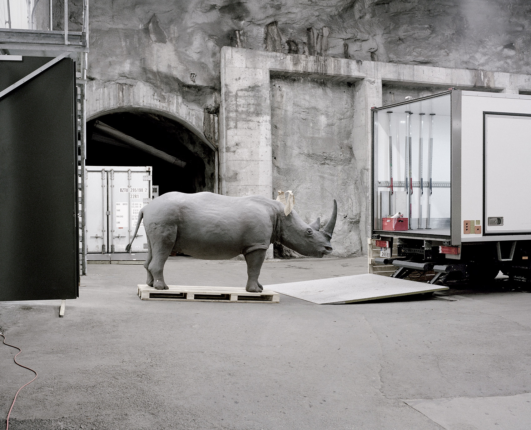 A Moveable Beast by Helge Skodvin — A taxidermied rhinoceros stands on a wooden pallet platform in an industrial storage facility in Bergen, Norway.
