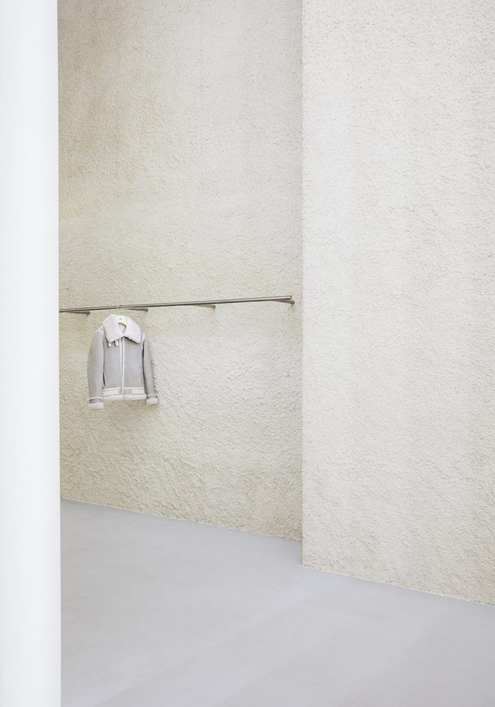 J. Lindeberg by Studio David Thulstrup — A single grey zip-up jacket hangs from a horizontal steel rod mounted at mid-wall height, creating a minimal display against textured render walls in warm...