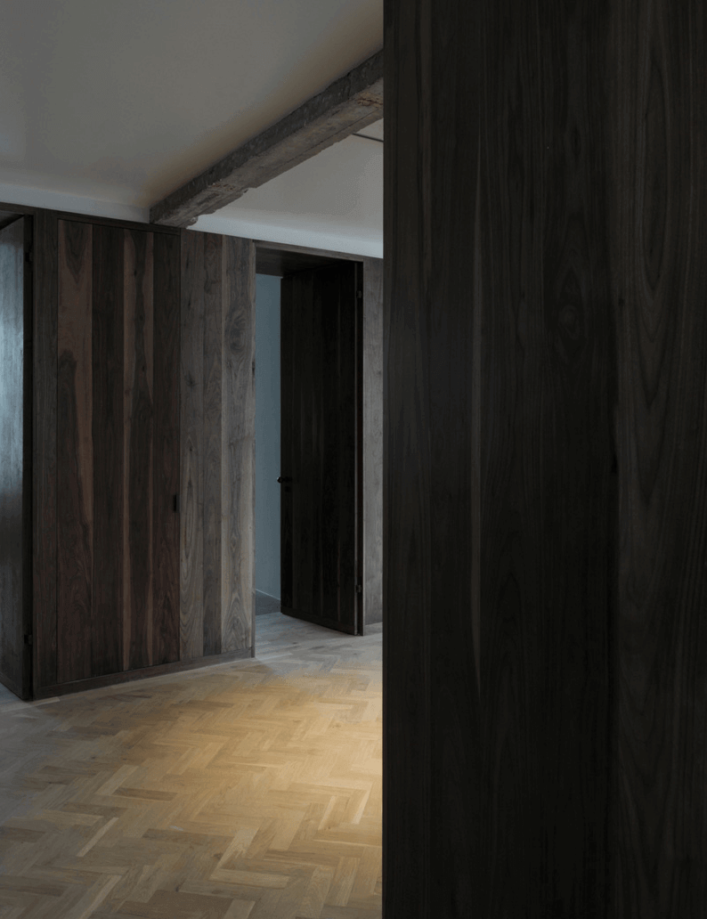 Queens Court by Feilden Fowles — A corridor in Queens Court frames an axial perspective through dark walnut wall paneling that extends floor to ceiling on both sides, creating a linear...