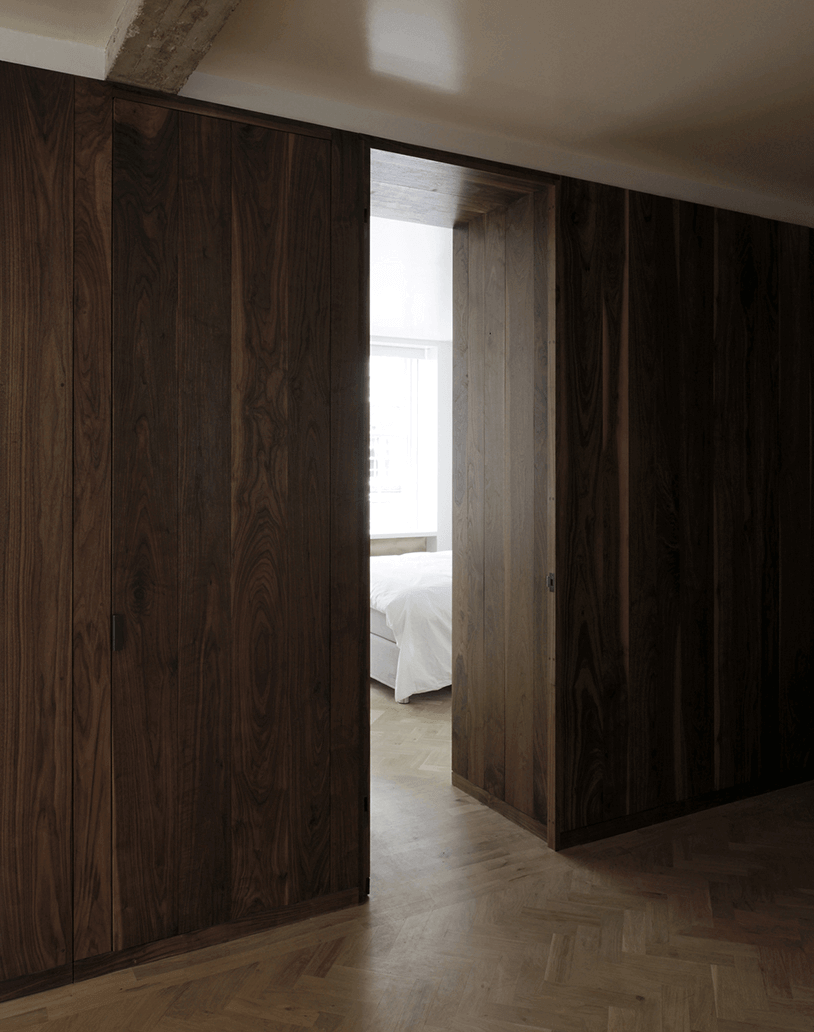 Queens Court by Feilden Fowles — A symmetrical corridor passage framed by dark walnut wall paneling that extends floor to ceiling on both sides, creating a narrow volumetric enclosure.
