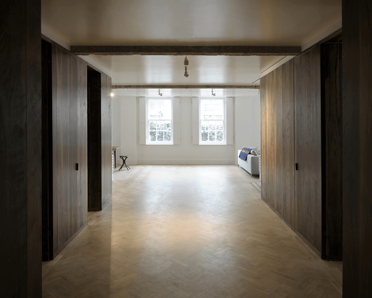 Queens Court by Feilden Fowles — A corridor framed by symmetrical dark walnut wall paneling extending floor to ceiling, creating a pronounced linear perspective toward a distant threshold.