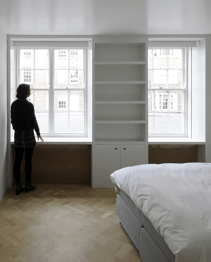 Queens Court by Feilden Fowles — A bedroom in Queens Court features a symmetrical composition anchored by built-in cabinetry between two tall sash windows.