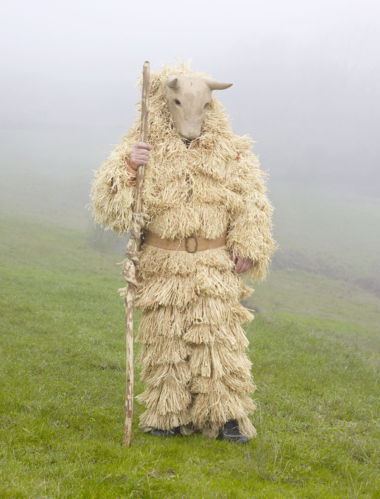 Wilder Mann by Charles Fréger — A figure stands centrally in a fog-laden pastoral landscape, fully encased in overlapping rows of bleached straw and dried plant fibers that extend from...