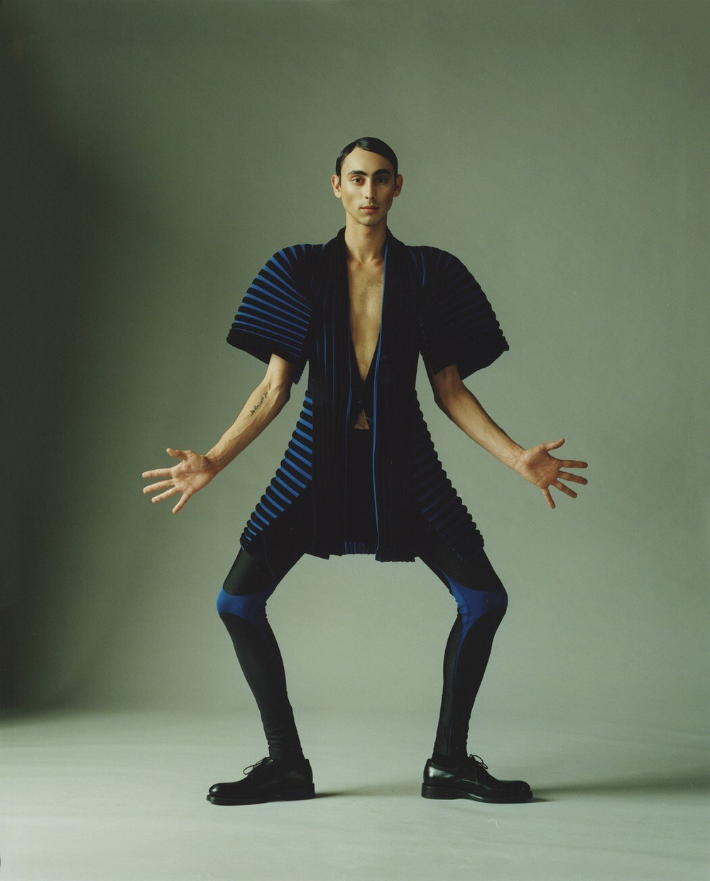MA Collection by Martin Tual by Martin Taul — A model wearing Martin Tual's MA Collection strikes a wide-legged stance against a sage-green backdrop.