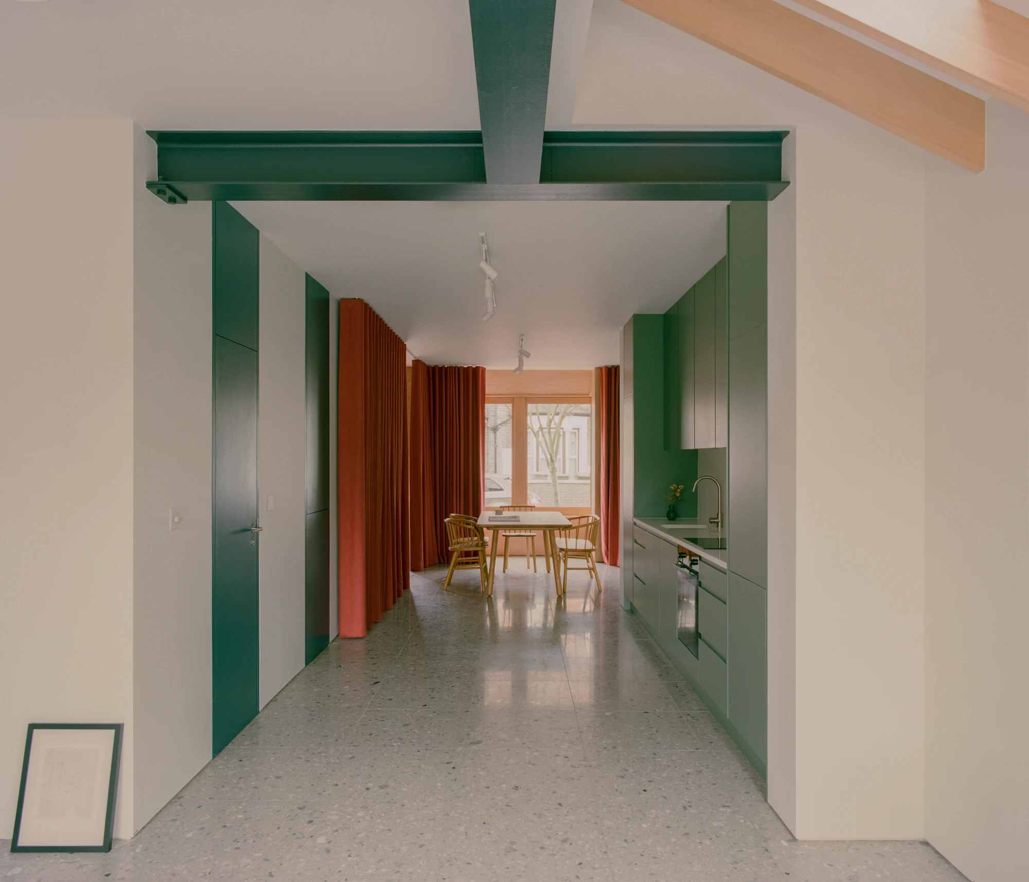 Walthamstow House by PSL — A galley-style kitchen corridor lined with deep forest-green cabinetry on both sides, extending toward a dining area framed by rust-orange curtains.