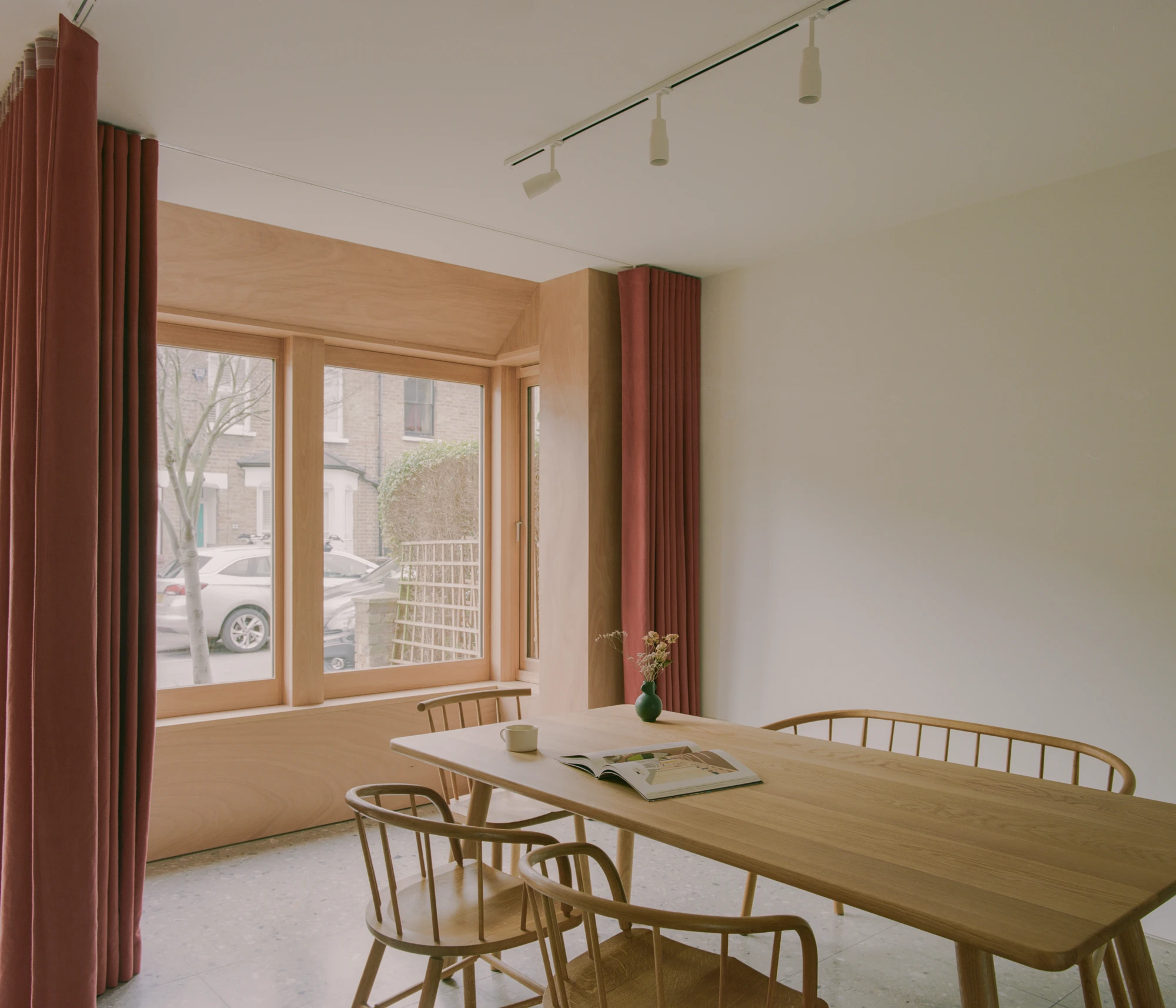 Walthamstow House by PSL — A dining space with oak table and spindle-back chairs positioned perpendicular to a bay window, framing a street view of parked cars and brick facades.