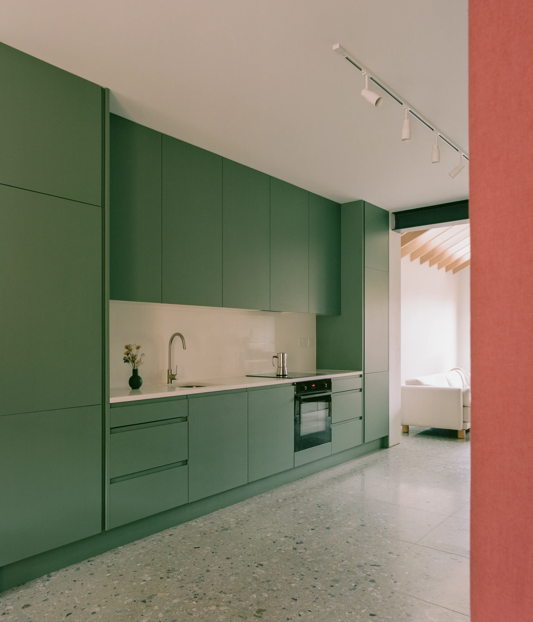 Walthamstow House by PSL — The kitchen occupies a single wall in an open-plan ground floor, defined by recessed cabinetry in deep sage-green with a cream-colored tile backsplash.