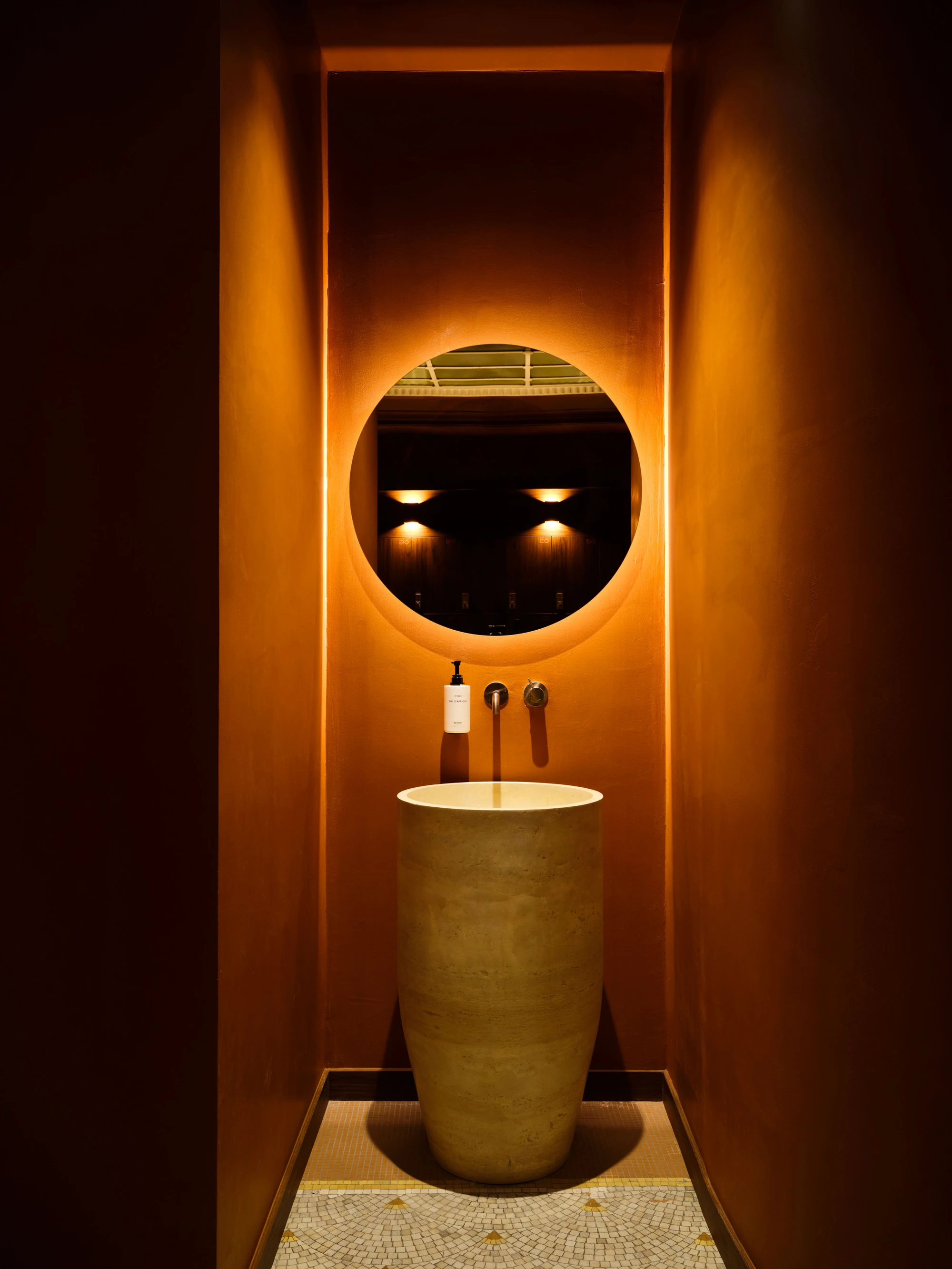 Sant Roch Sauna by Futurstudio — A cylindrical stone vessel sink anchors this narrow bathroom passage, positioned beneath a large circular backlit mirror that casts warm orange light across...