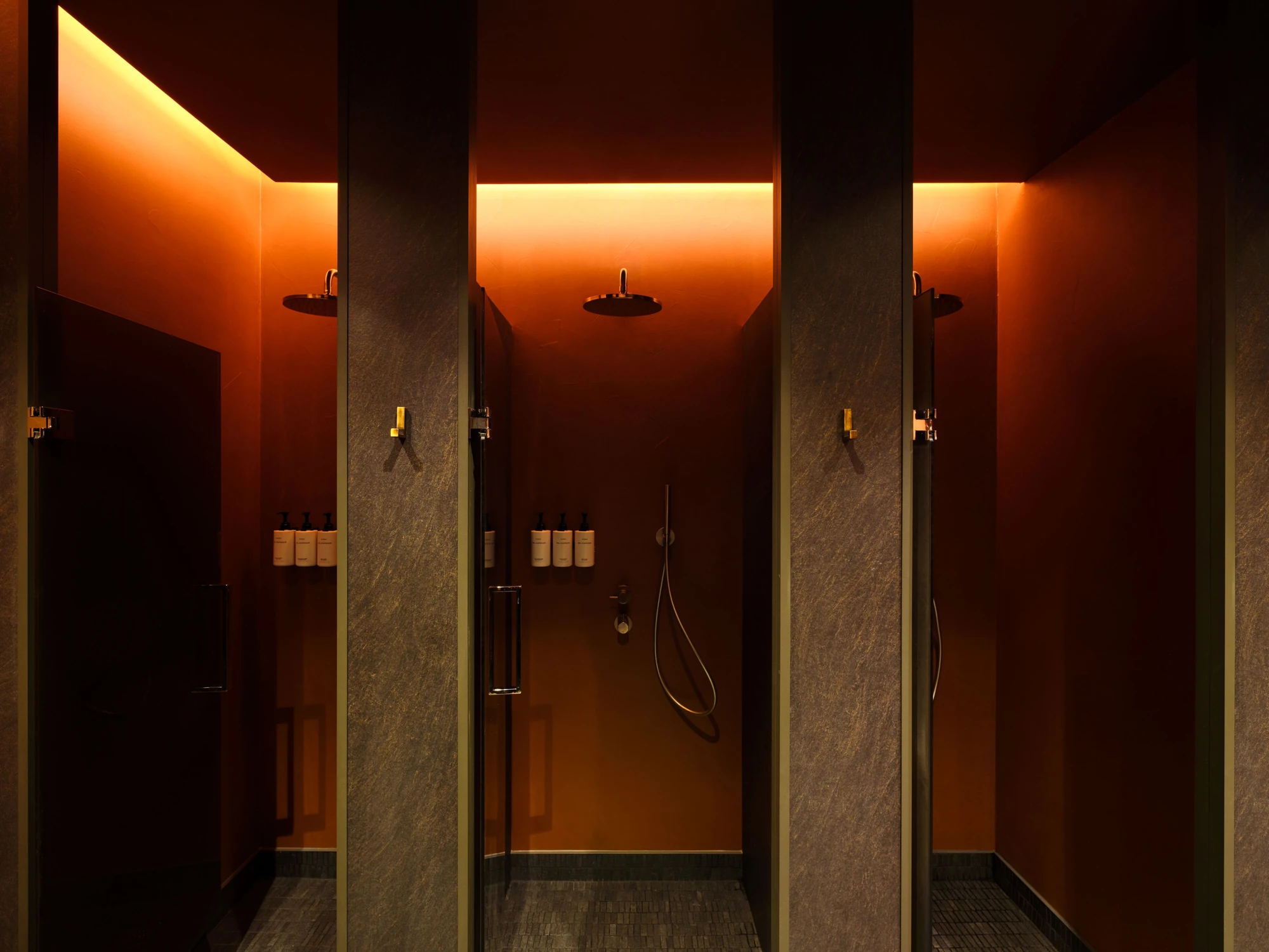 Sant Roch Sauna by Futurstudio — Three shower cubicles separated by textured stone partitions display backlit orange-red walls creating volumetric depth.