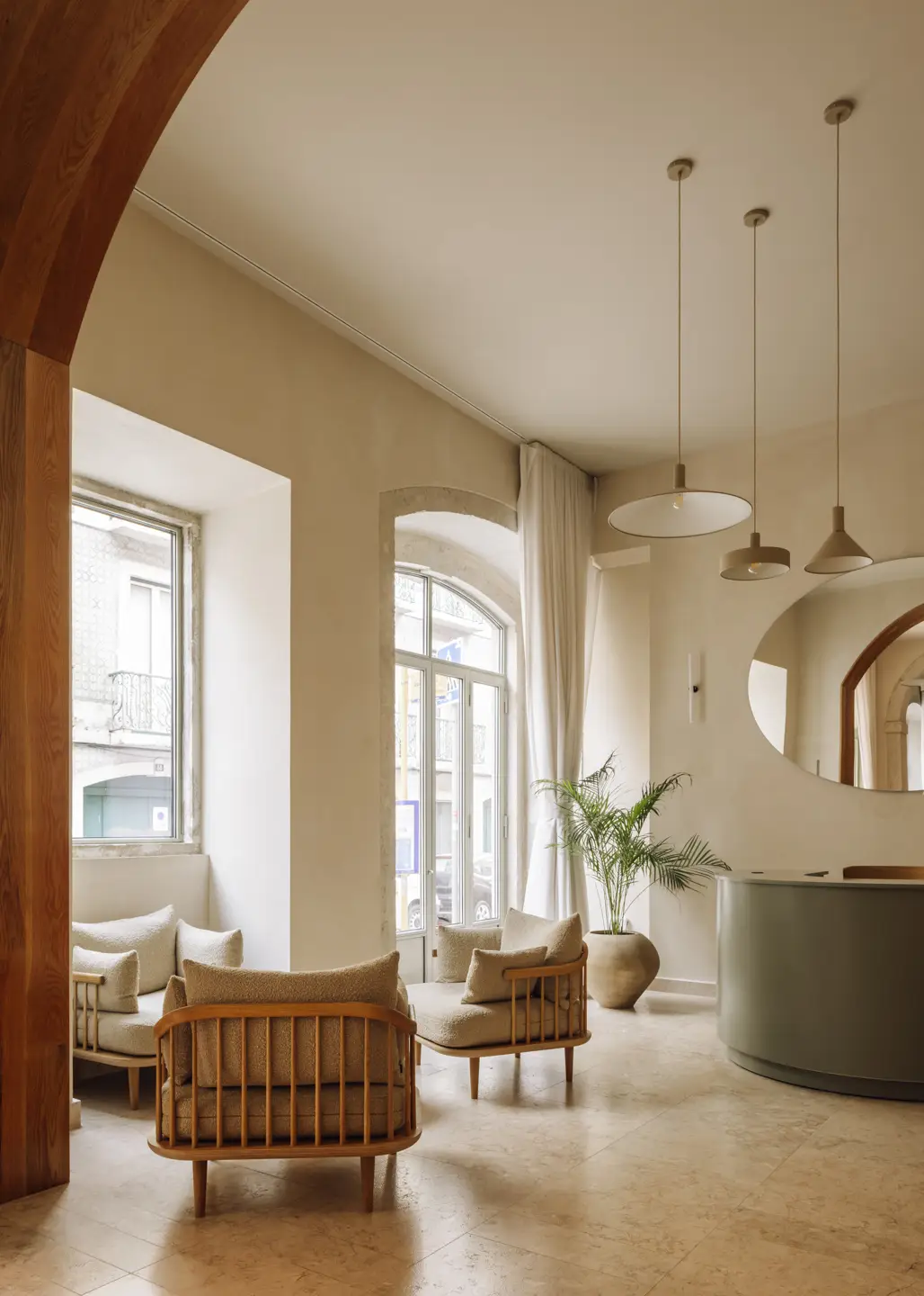 The Verse by João Luís Carrilho da Graça + Studio Astolfi — A reception or common area bathed in natural light from tall arched windows, furnished with mid-century wooden lounge chairs upholstered in cream linen.