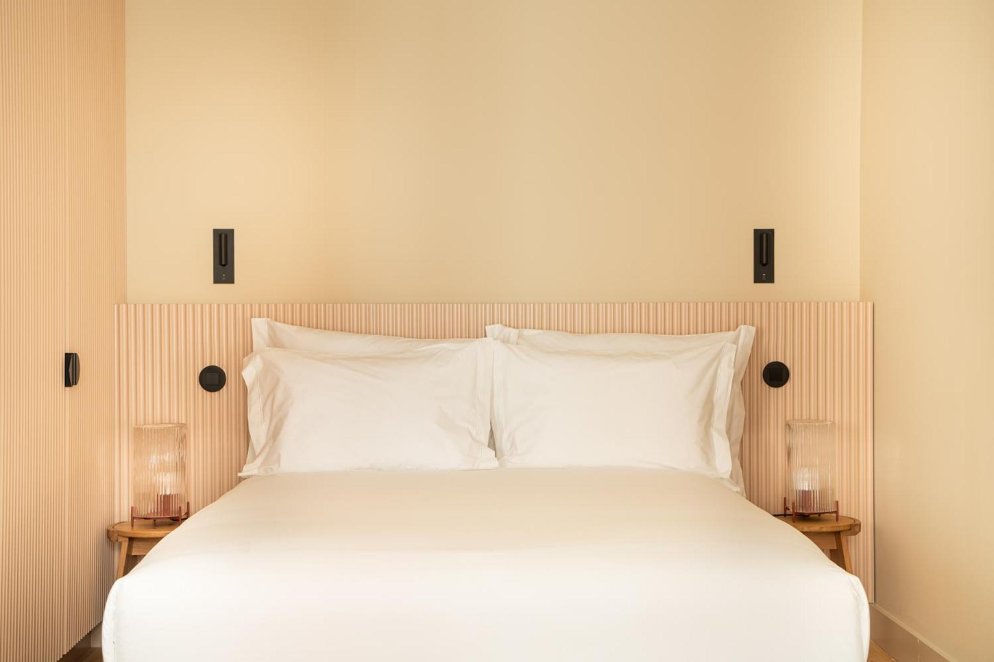 The Verse by João Luís Carrilho da Graça + Studio Astolfi — A bedroom interior centered on a bed with cream linen bedding and white pillows set against a headboard clad in vertical timber slats in light natural finish.