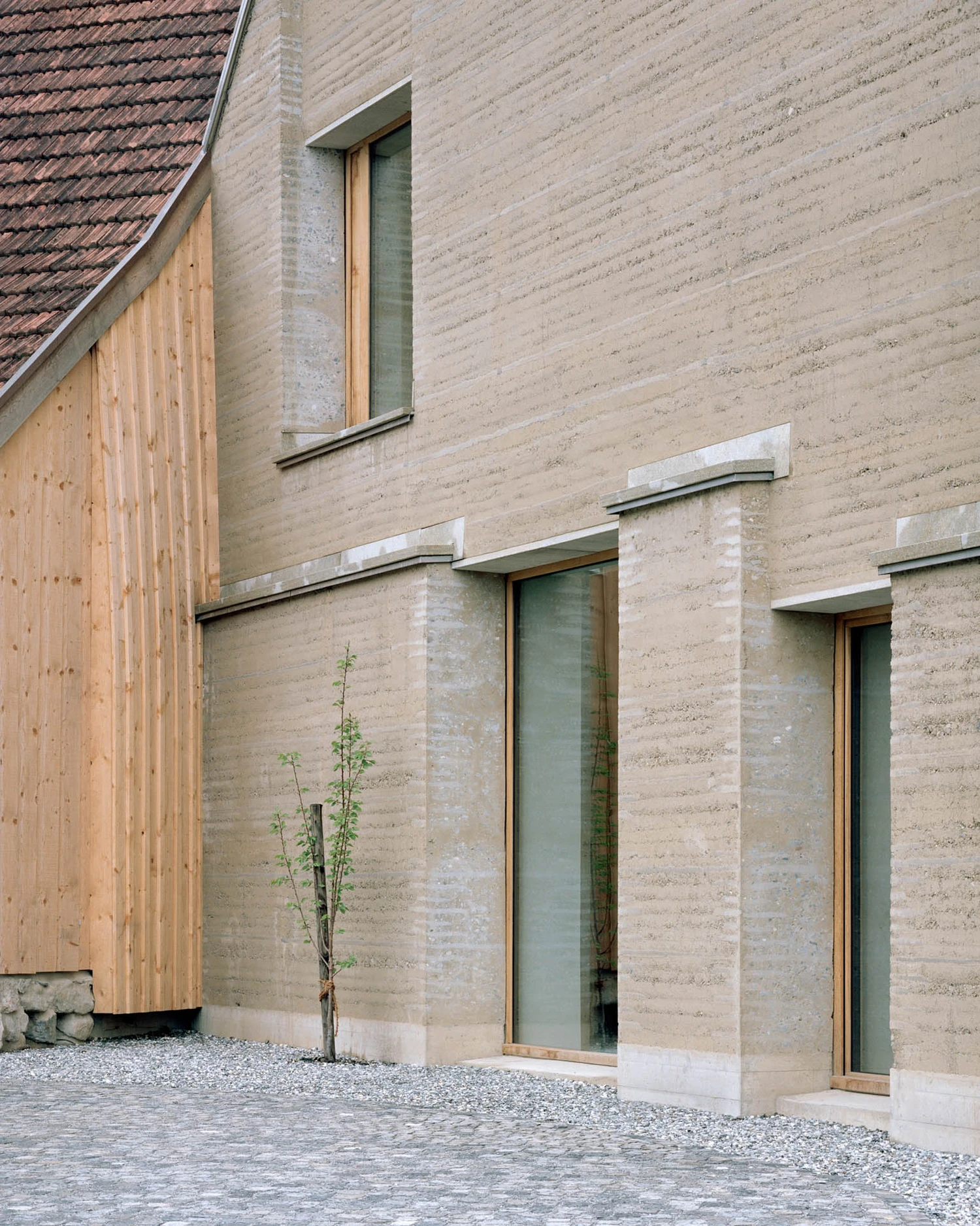 The Gässli5 by allen + crippa architektur — The rammed-earth facade displays pronounced horizontal stratification across its matte surface, with timber-framed windows set flush into the brick plane...