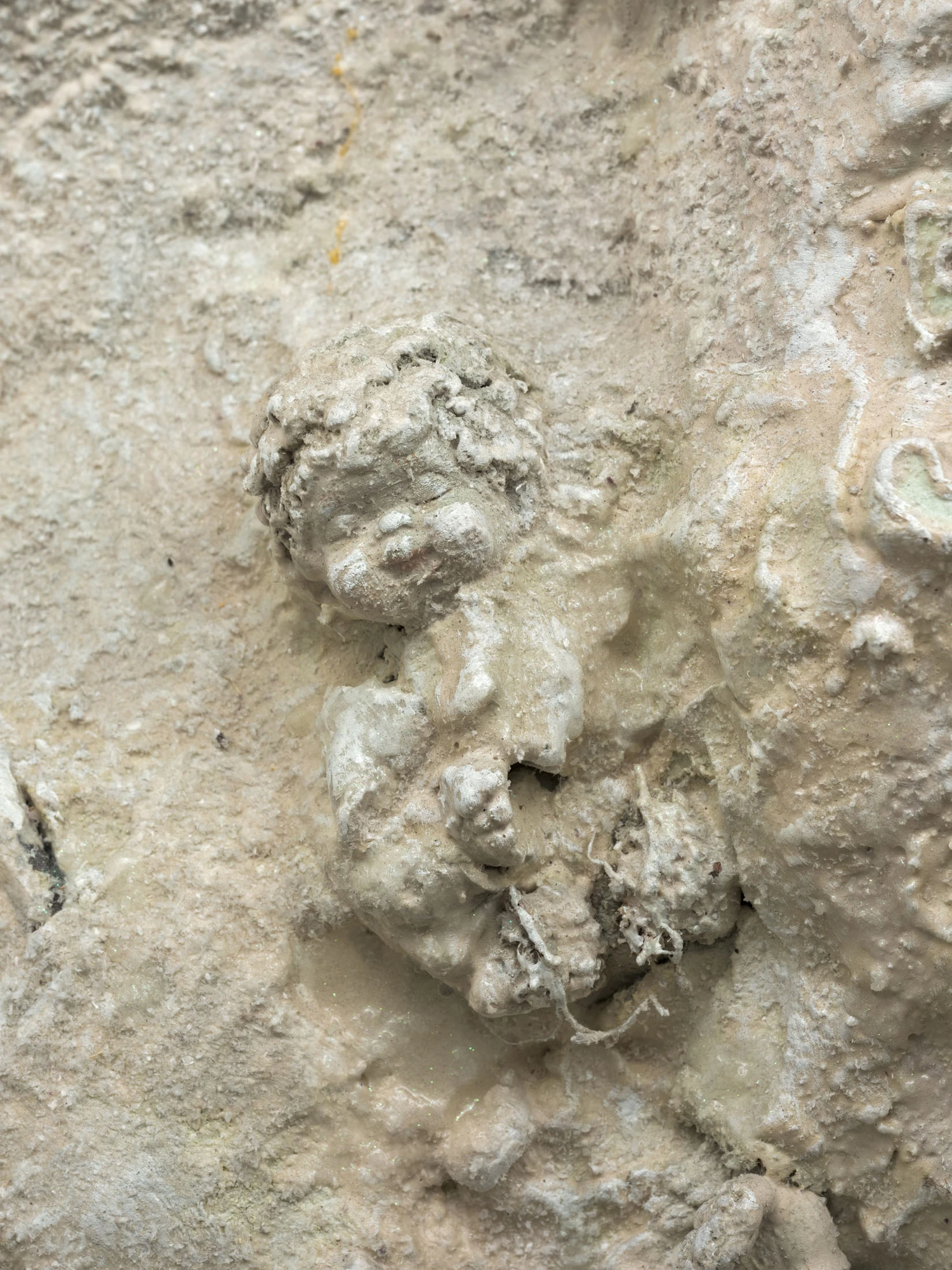 Cold Front by Trey Abdella at Kraupa-Tuskany Zeidler — A relief-carved stone or plaster cherub head occupies the center of this close-up composition, surrounded by granular cream-colored surface material.