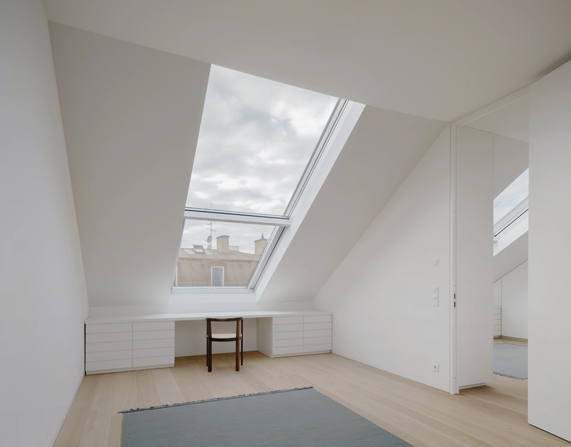 Ursulastraße 6 by Studio Mark Randel — A top-floor attic room in Munich's Altschwabing features a large rectangular skylight that floods the space with daylight, revealing a pitched white ceiling...