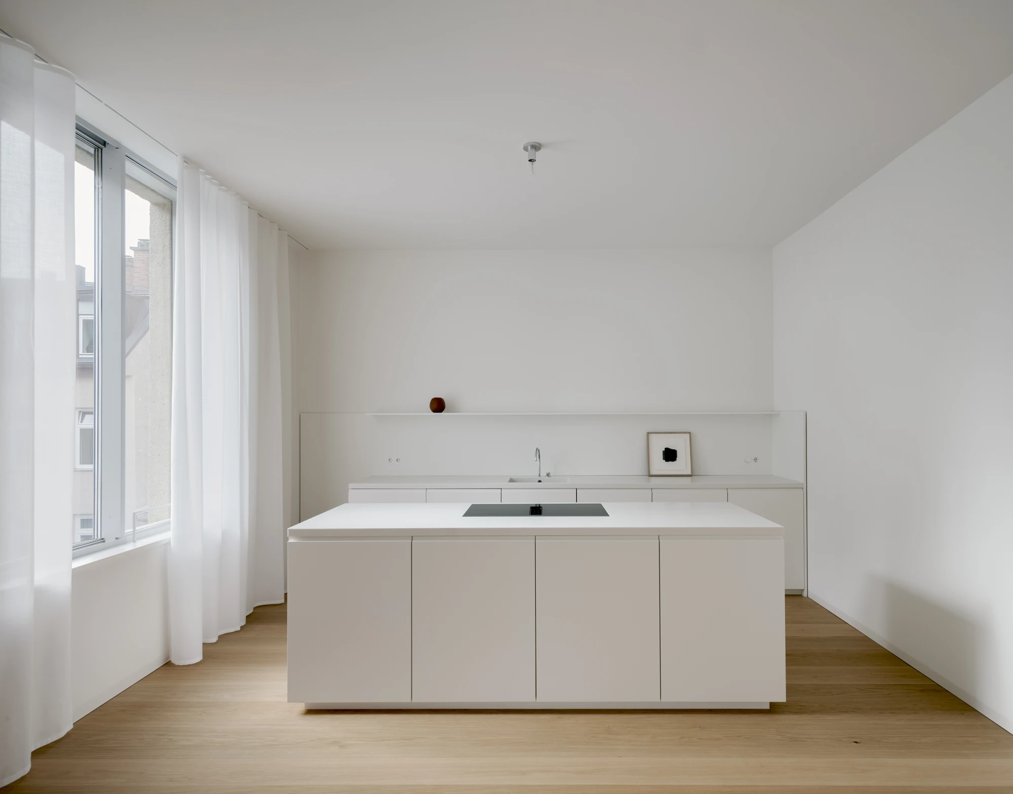 Ursulastraße 6 by Studio Mark Randel — A minimalist kitchen island occupies the center of a white-walled interior, its clean-lined cabinetry faced in white lacquered panels with recessed hardware.