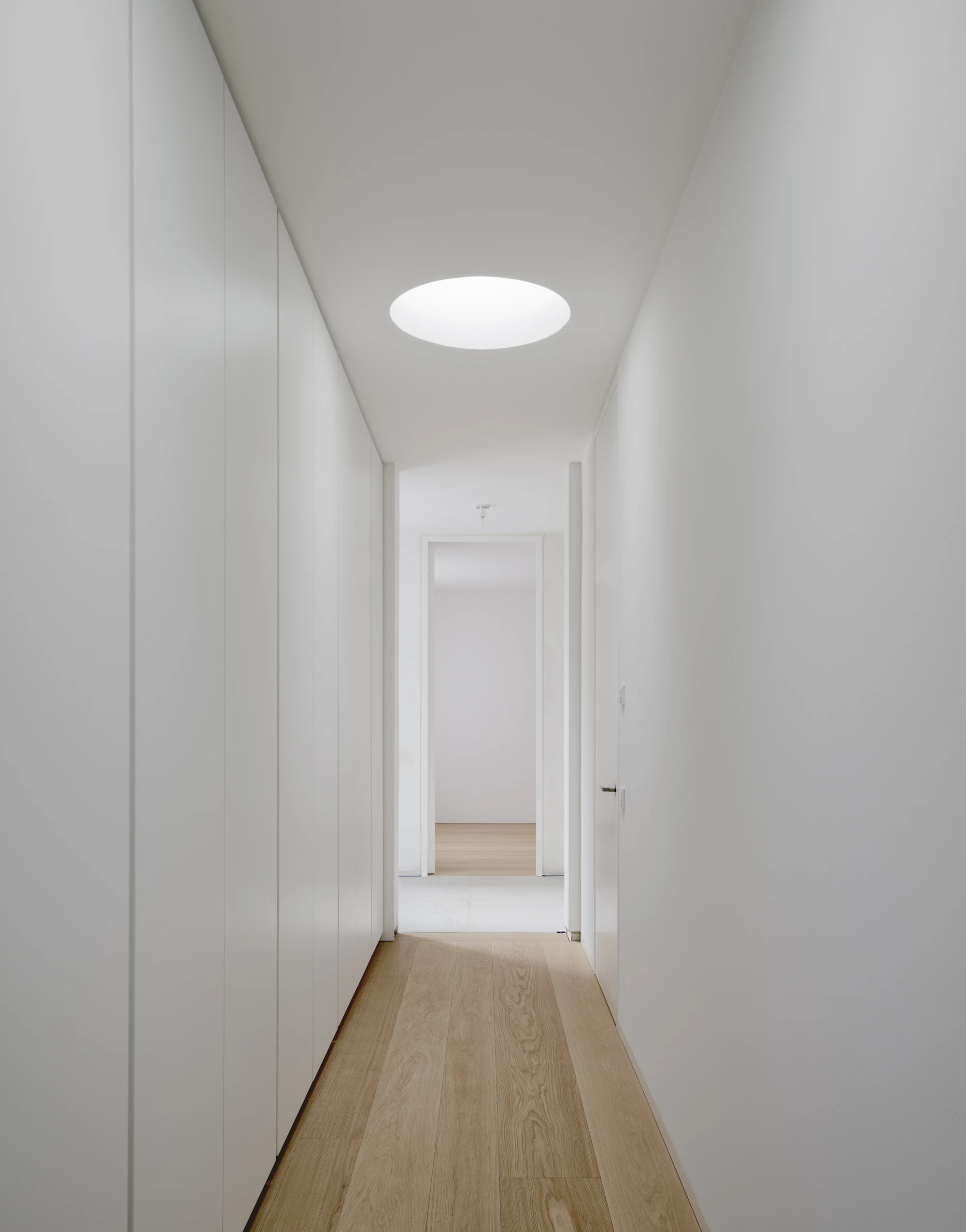 Ursulastraße 6 by Studio Mark Randel — A corridor stretches toward multiple doorways in receding perspective, lined with white-painted walls and timber flooring in light oak tones.
