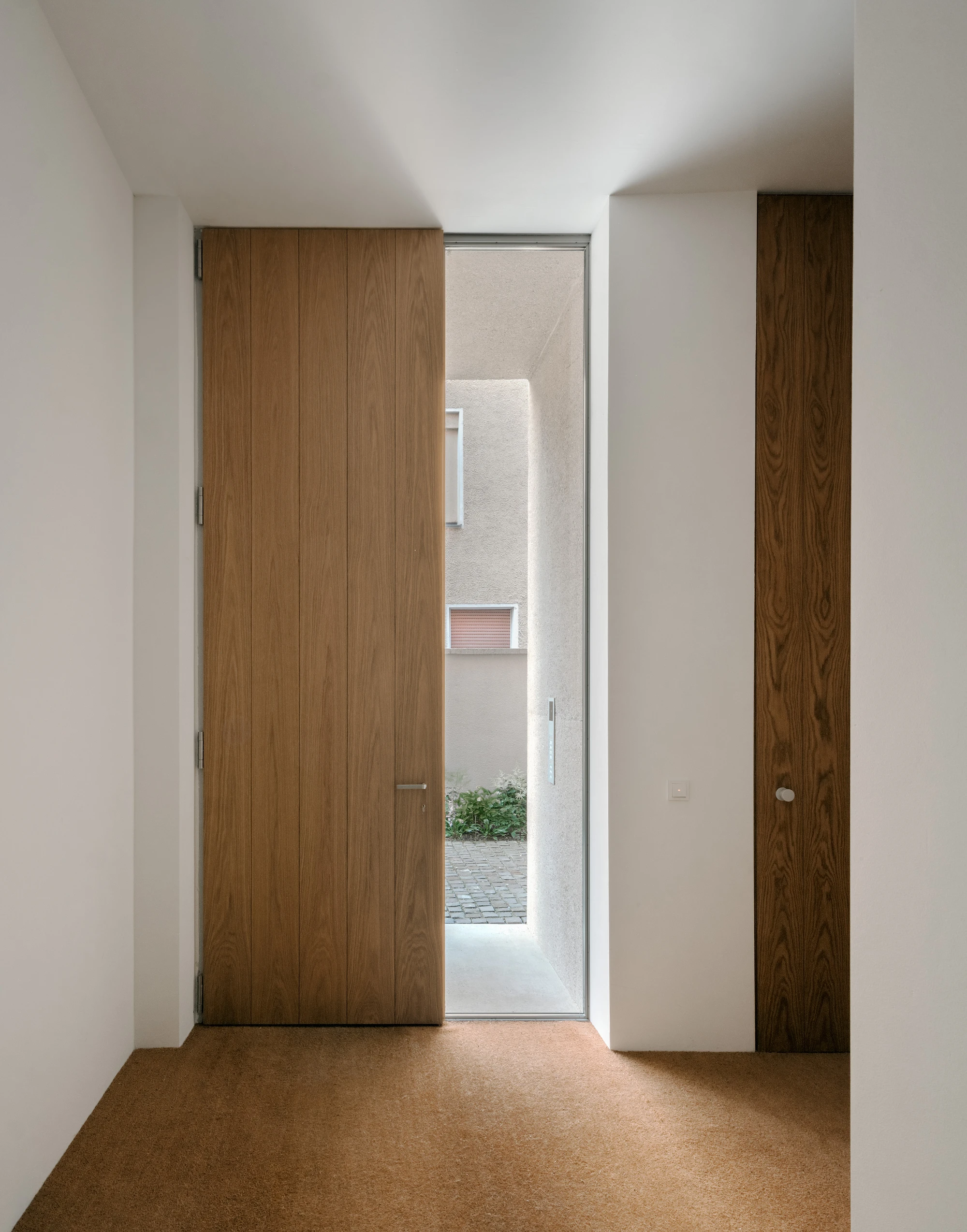 Ursulastraße 6 by Studio Mark Randel — A passage frames the threshold between interior and exterior through a timber-clad sliding door with minimal aluminum framing.