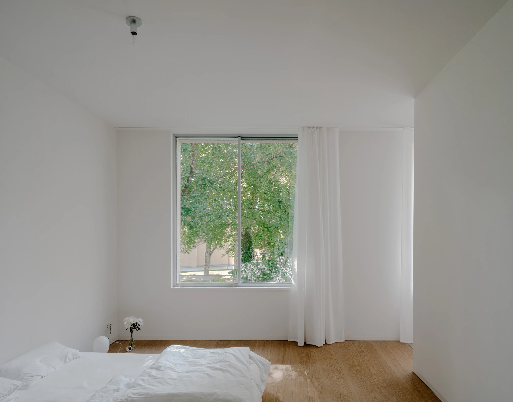 Ursulastraße 6 by Studio Mark Randel — A bedroom interior framed from the bed looking toward a large window with aluminum mullions revealing a mature tree canopy beyond.