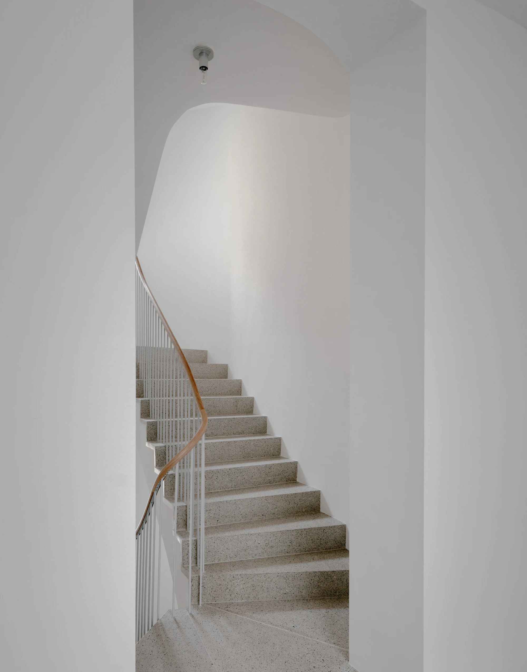 Ursulastraße 6 by Studio Mark Randel — A stairwell ascends through a volumetric white interior, its concrete treads anchored by a minimal white metal balustrade and warm copper handrail that...