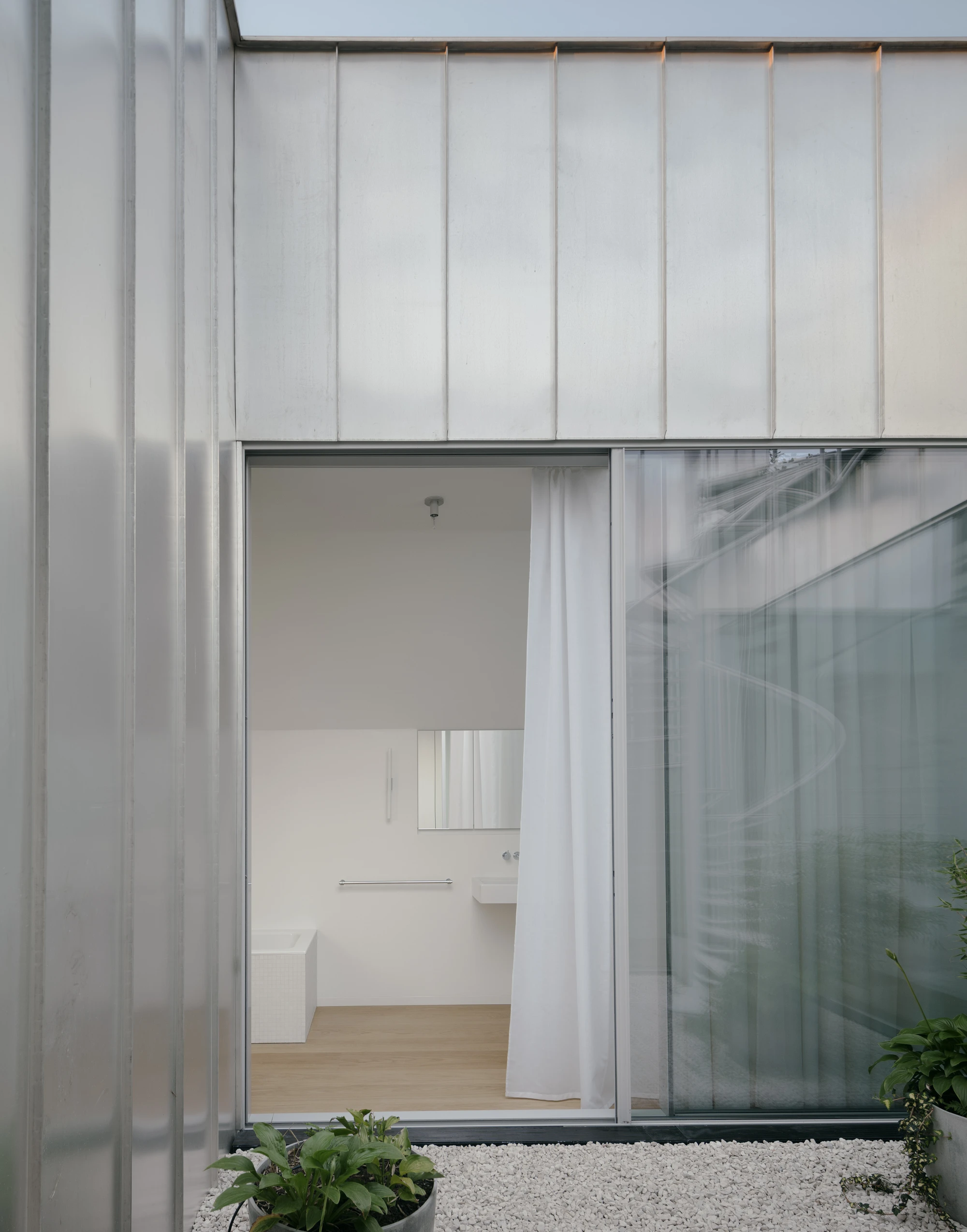 Ursulastraße 6 by Studio Mark Randel — A frameless glass sliding door opens onto a minimalist interior, its threshold marked by a transition from exterior gravel to wooden floorboards.