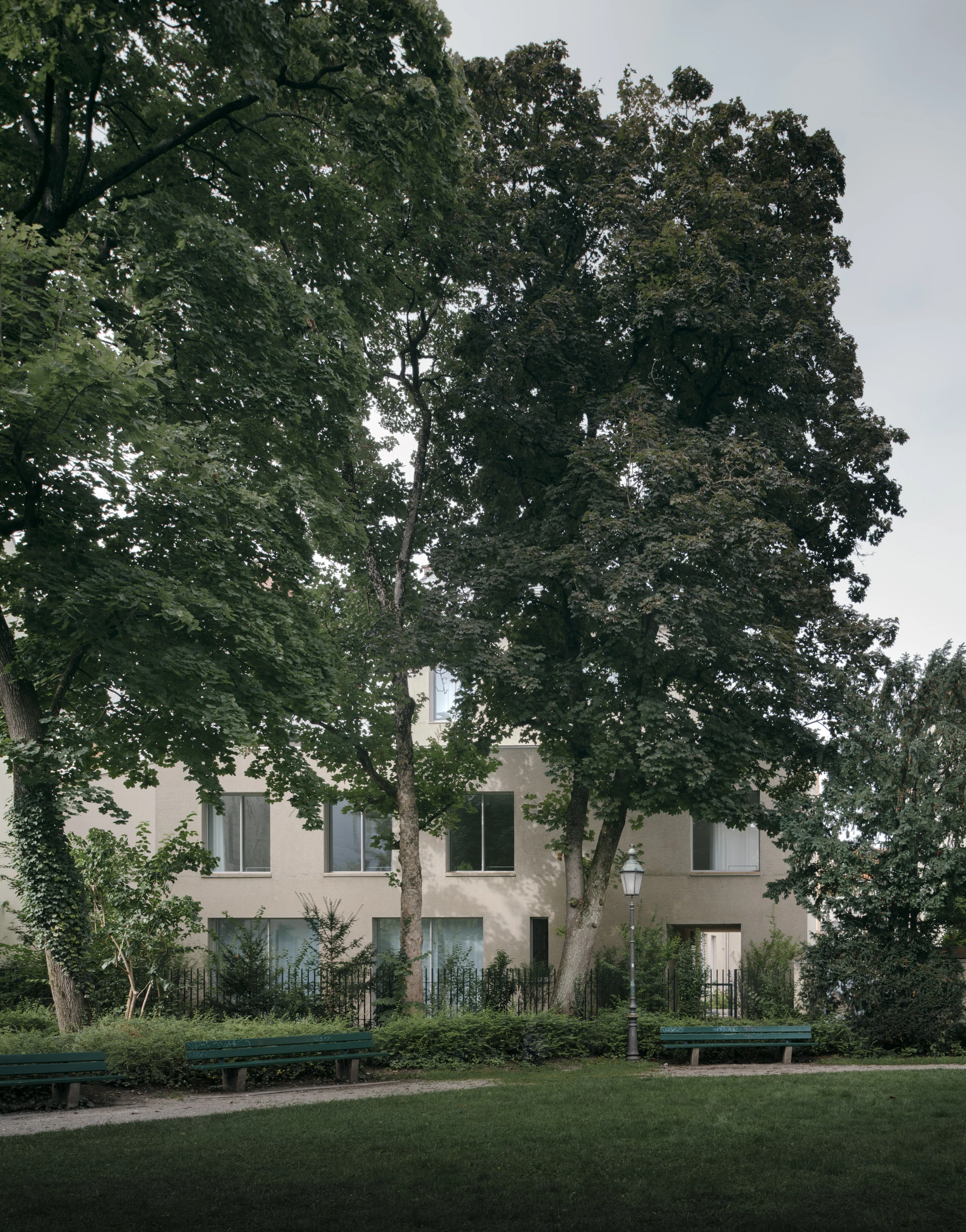 Ursulastraße 6 by Studio Mark Randel — Mature deciduous trees frame a rendered facade in a Munich park setting, their dense canopies creating dappled light across the cream-coloured surface.