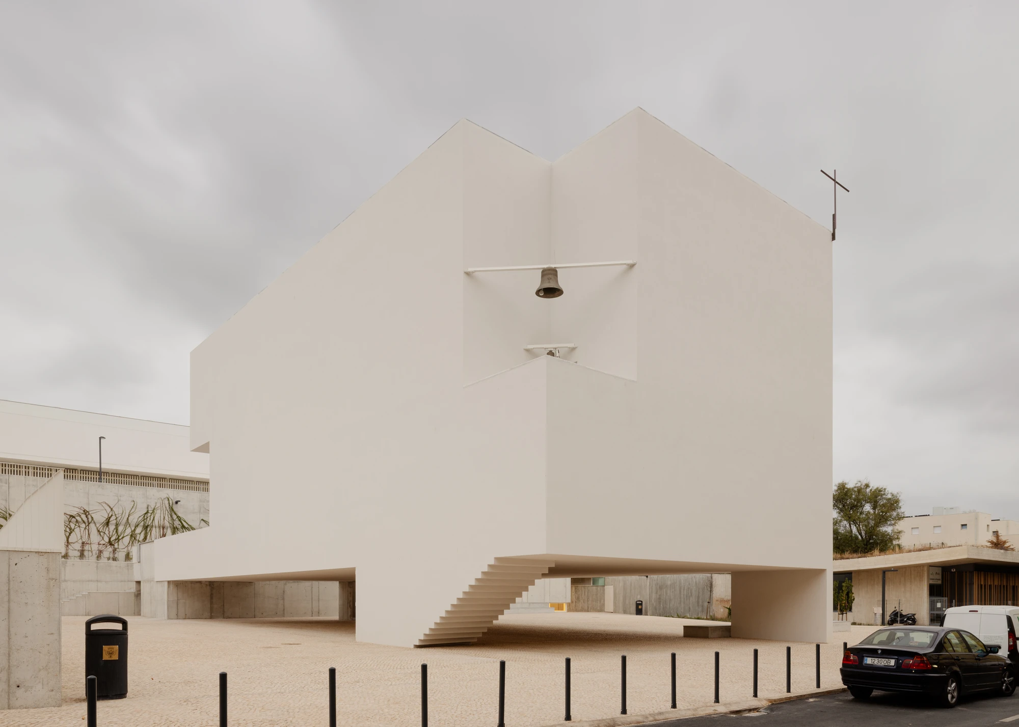 Da Boavista Church by Matos Gameiro arquitectos + atelier bugio — Da Boavista Church presents a monumental rendered facade facing a paved plaza, its geometric form defined by two pitched roof volumes meeting at a central...