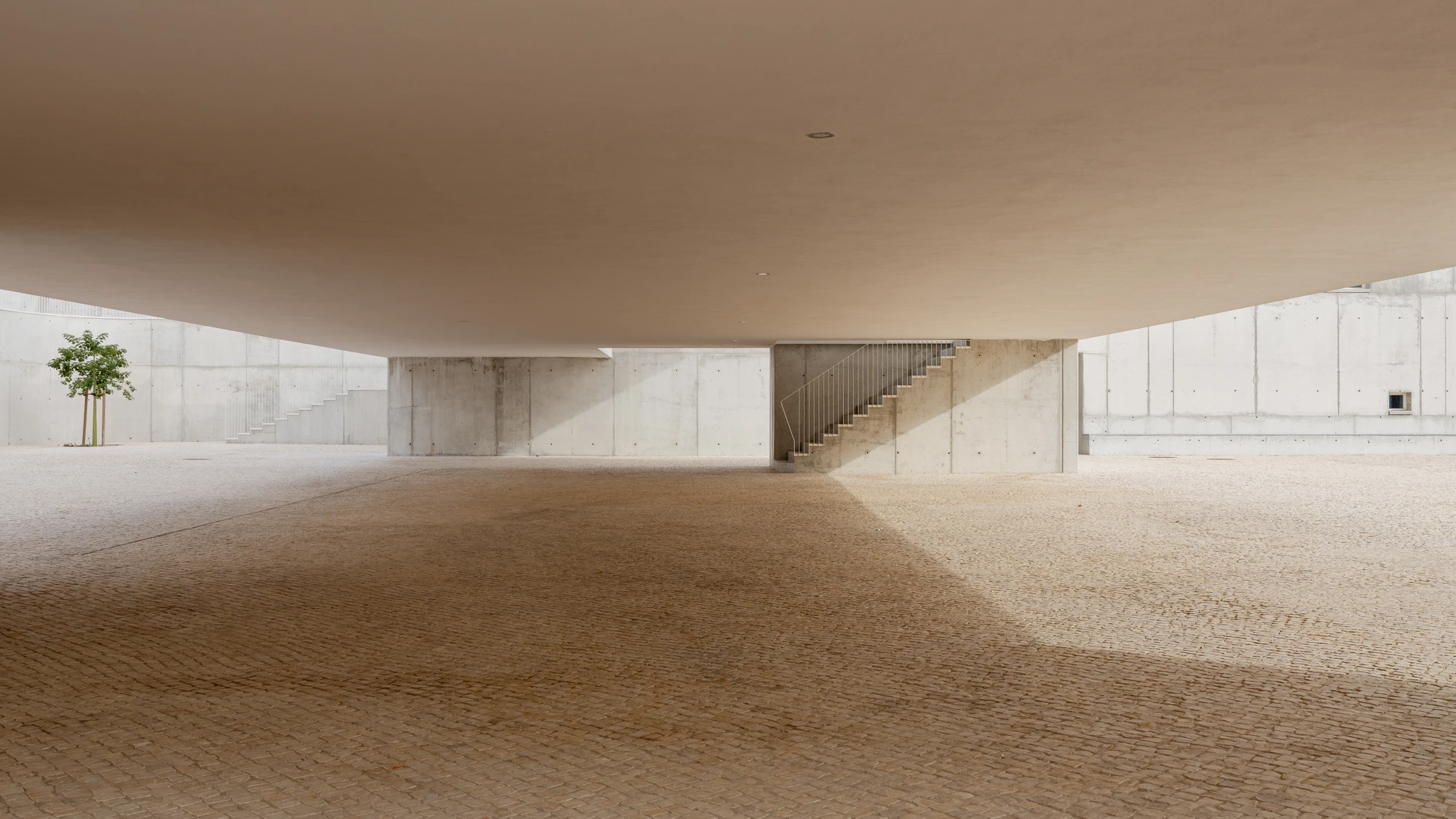 Da Boavista Church by Matos Gameiro arquitectos + atelier bugio — A rendered concrete wall anchors the eastern edge of a public plaza in Lisbon, its monolithic surface punctuated by recessed staircase cutouts that create...