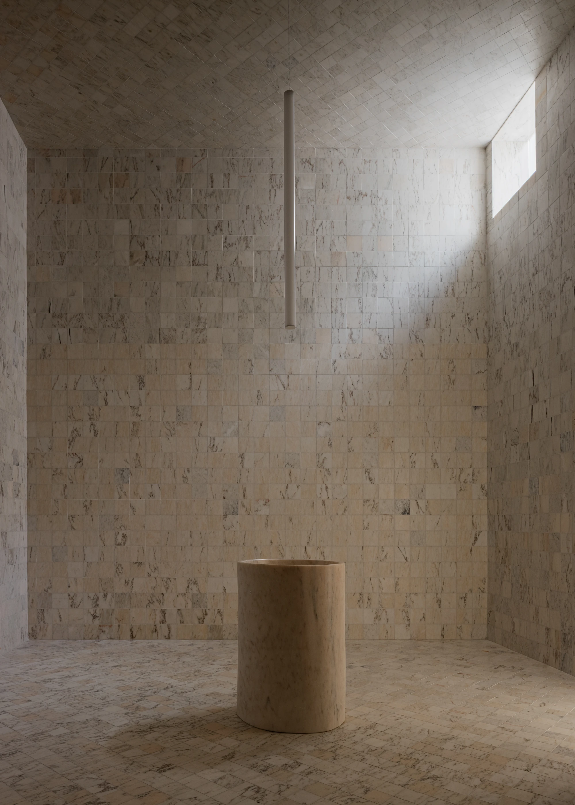 Da Boavista Church by Matos Gameiro arquitectos + atelier bugio — A monolithic travertine cylinder anchors the center of a rendered stone chamber, suspended beneath a single pendant steel rod descending from the coffered...