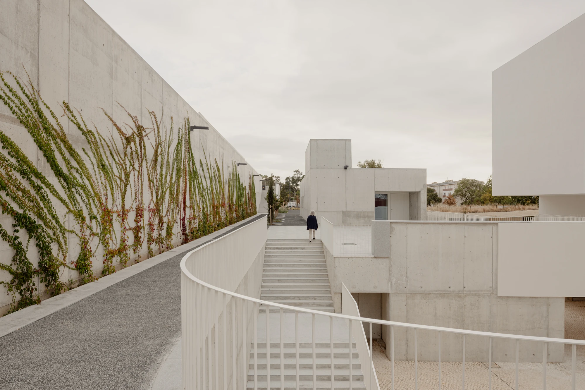 Da Boavista Church by Matos Gameiro arquitectos + atelier bugio — A curved concrete railing guides movement through a stepped plaza, its white-painted metal balustrade echoing the monolithic rendered wall that rises to the...