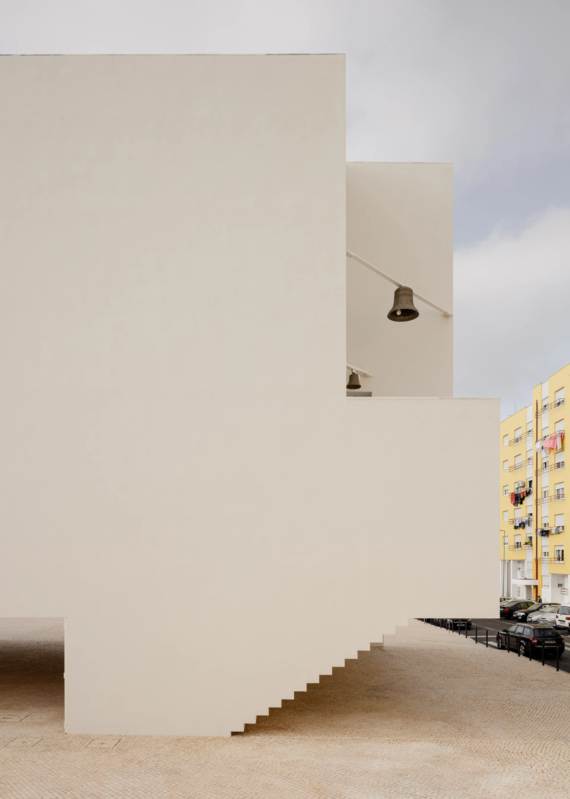 Da Boavista Church by Matos Gameiro arquitectos + atelier bugio — A monolithic rendered wall forms the primary architectural gesture, its cream-colored surface punctuated by two bronze bells suspended on angled steel brackets.