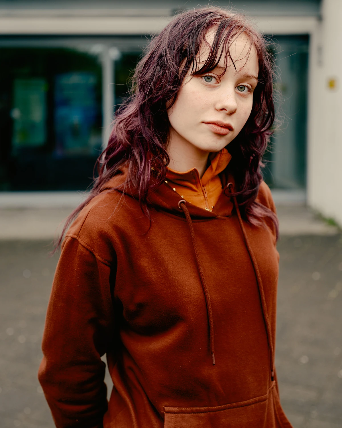 Lupine by Daniel Dorsa — Portrait of a young person with burgundy-dyed wavy hair, shot outdoors against a blurred institutional backdrop.
