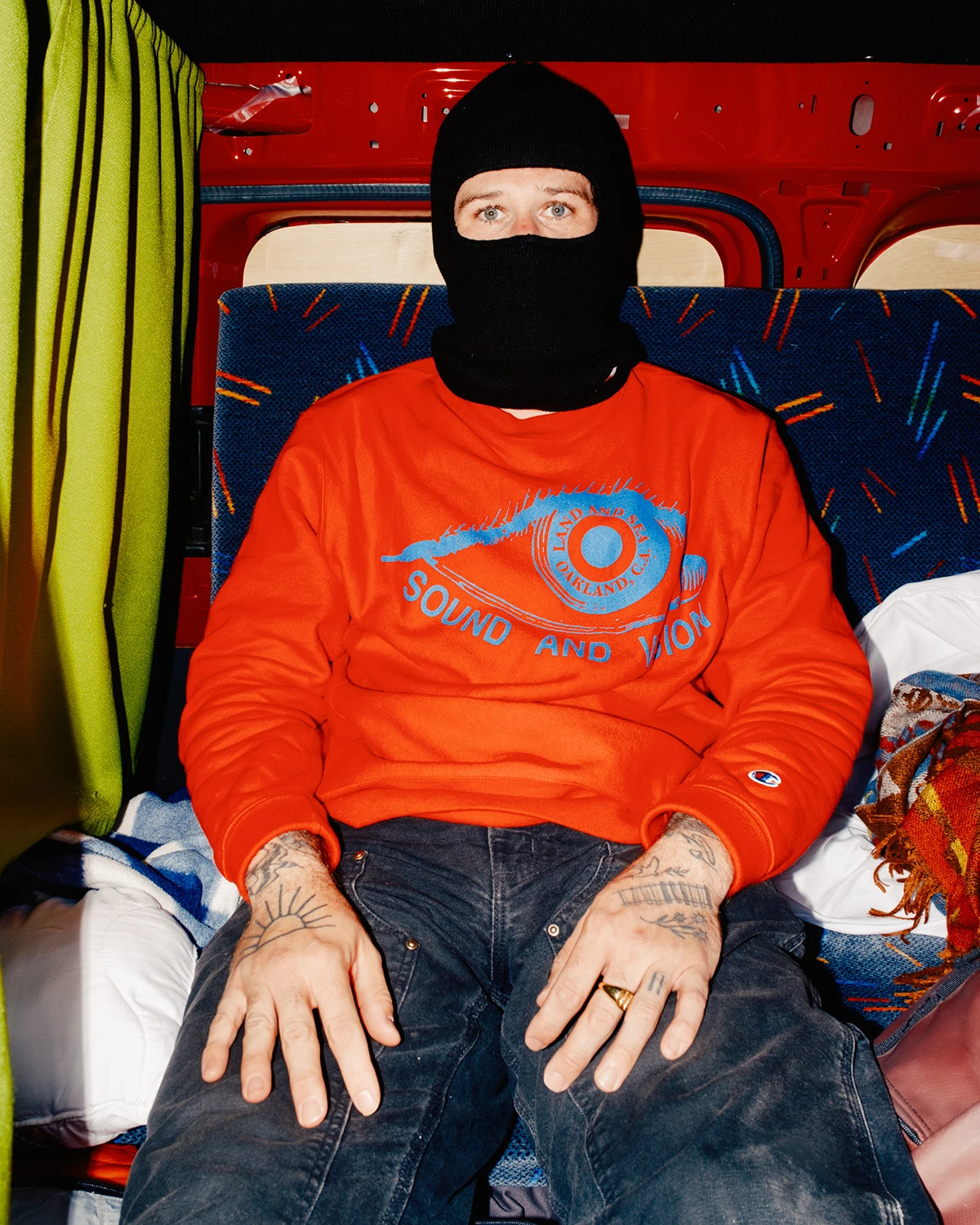 Lupine by Daniel Dorsa — A figure seated inside a coach interior wearing a black knit balaclava and bright orange crewneck sweatshirt printed with blue eye motif and 'SOUND AND...