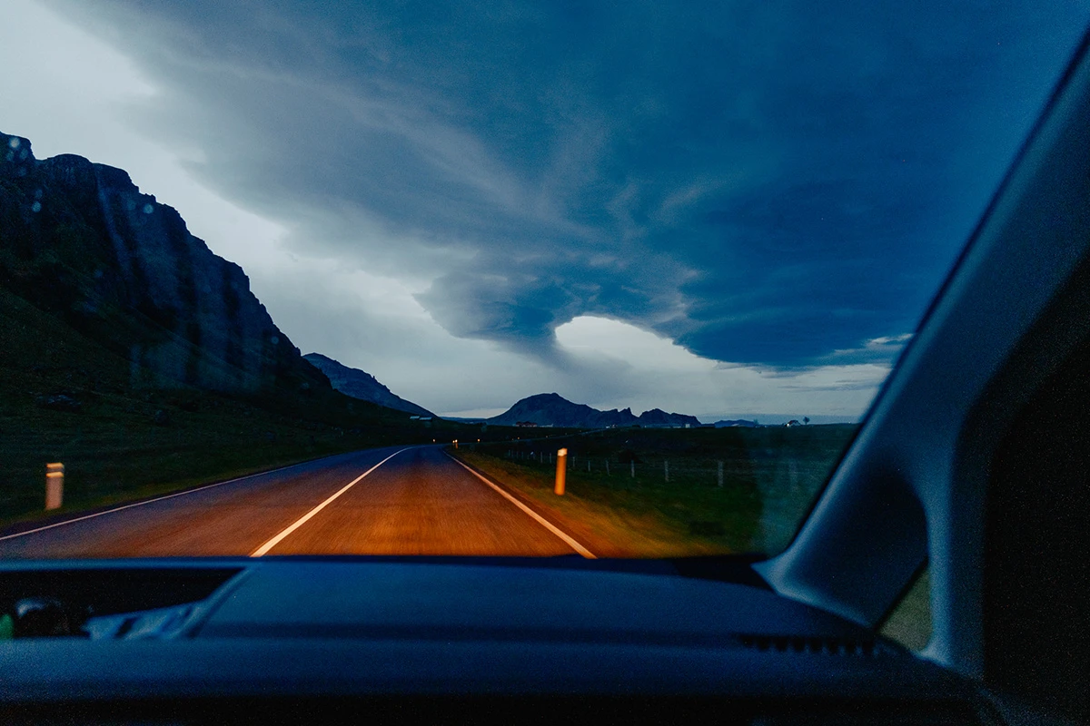 Lupine by Daniel Dorsa — Taken from inside a moving vehicle on an Icelandic highway at dusk, this photograph frames an illuminated asphalt road stretching toward distant volcanic peaks.