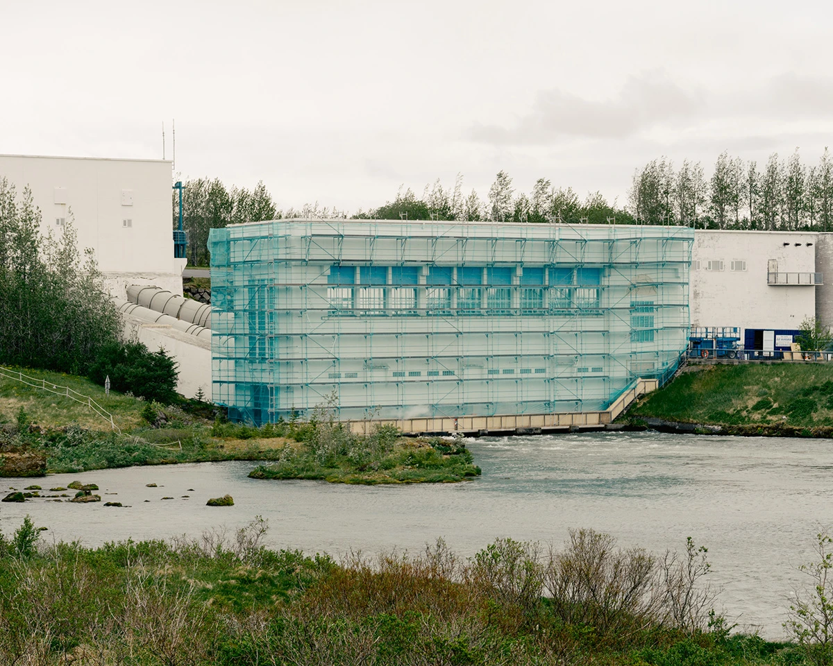 Lupine by Daniel Dorsa — A large-scale industrial structure wrapped in turquoise mesh scaffolding dominates the riverbank, its gridded facade suggesting ongoing construction or...