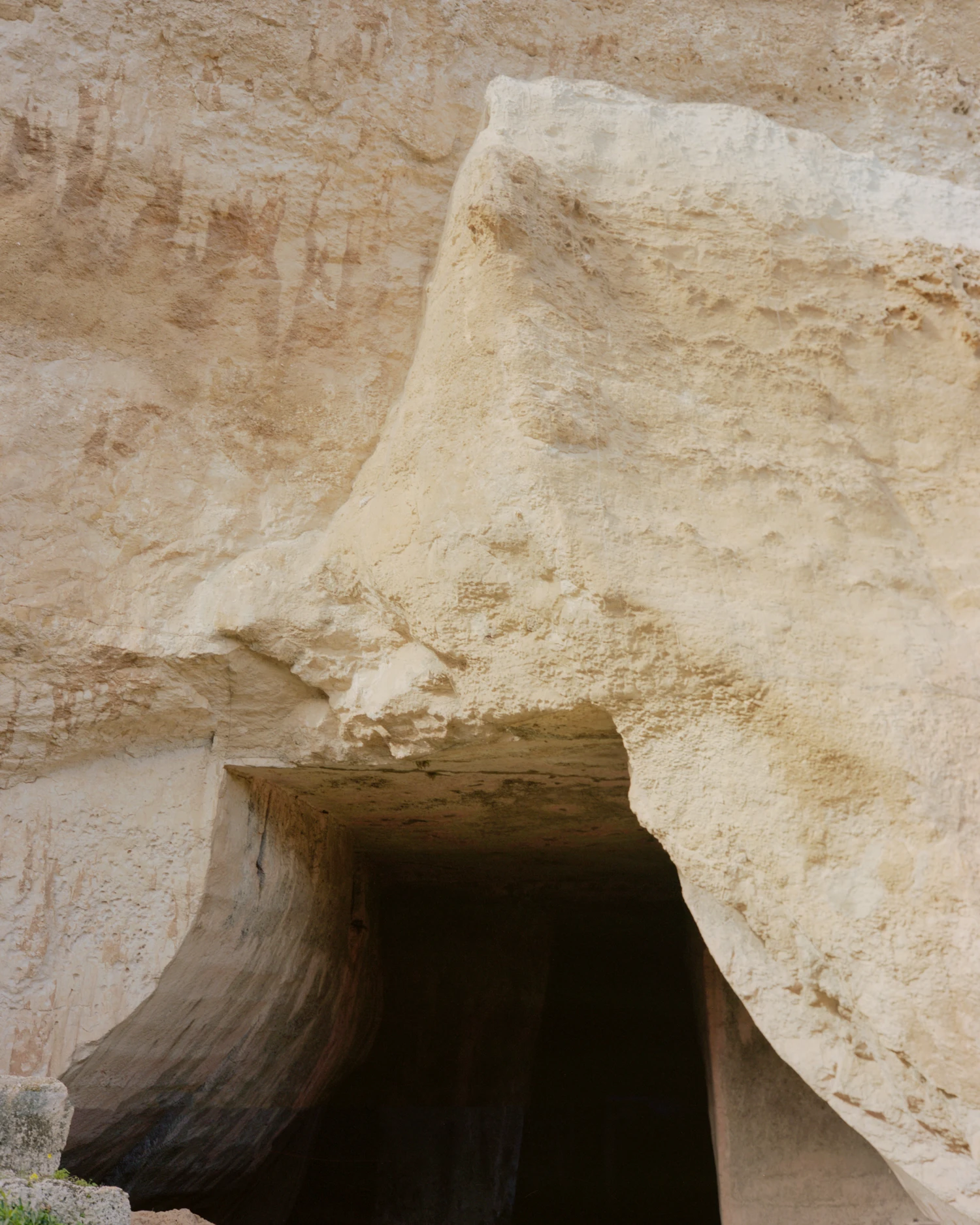 Pantalica Speculations by Leopold Banchini — Entrance to an underground chamber carved into cream-colored limestone, with a wooden timber lintel spanning the opening.