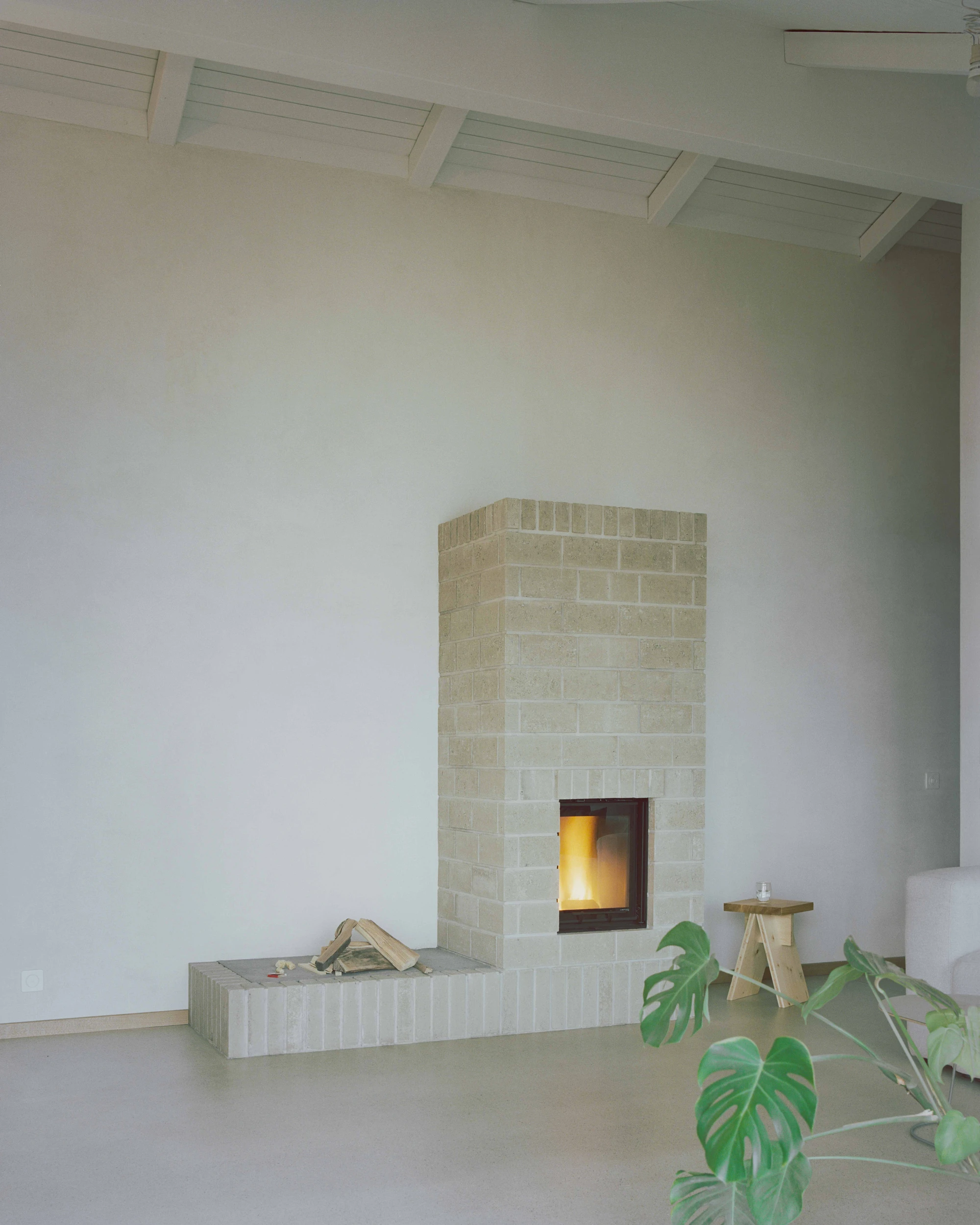 Family Villa by Noue Studio — A cream-brick fireplace rises monolithically from a white render base, its recessed opening framing an active fire.