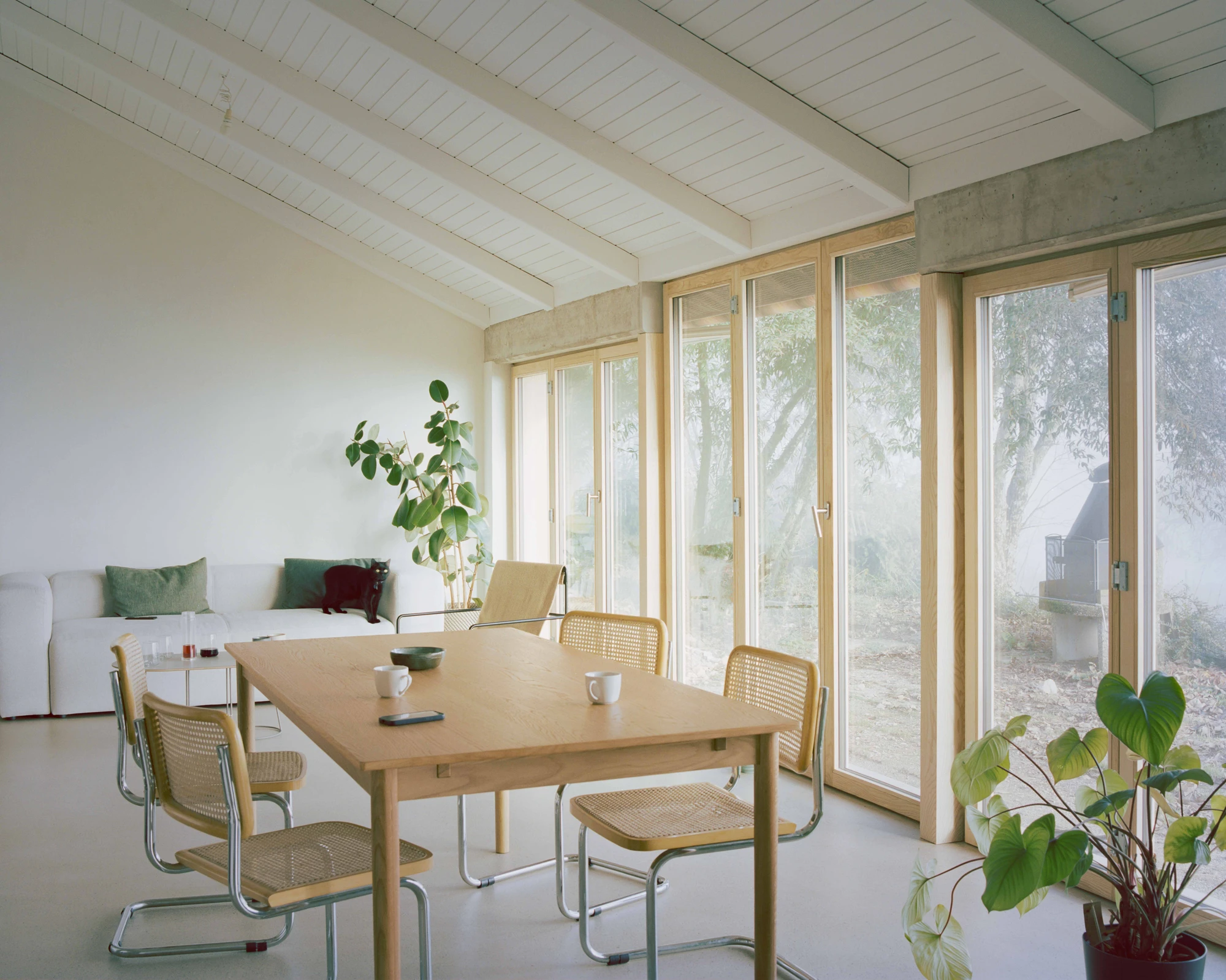 Family Villa by Noue Studio — A timber-framed dining space opens onto the landscape through a continuous run of bifold glass doors with natural wood casing.